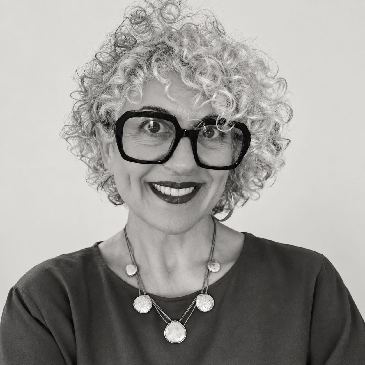 A smiling Laura wearing large black glasses, a dark top, and layered necklaces, against a plain background.
