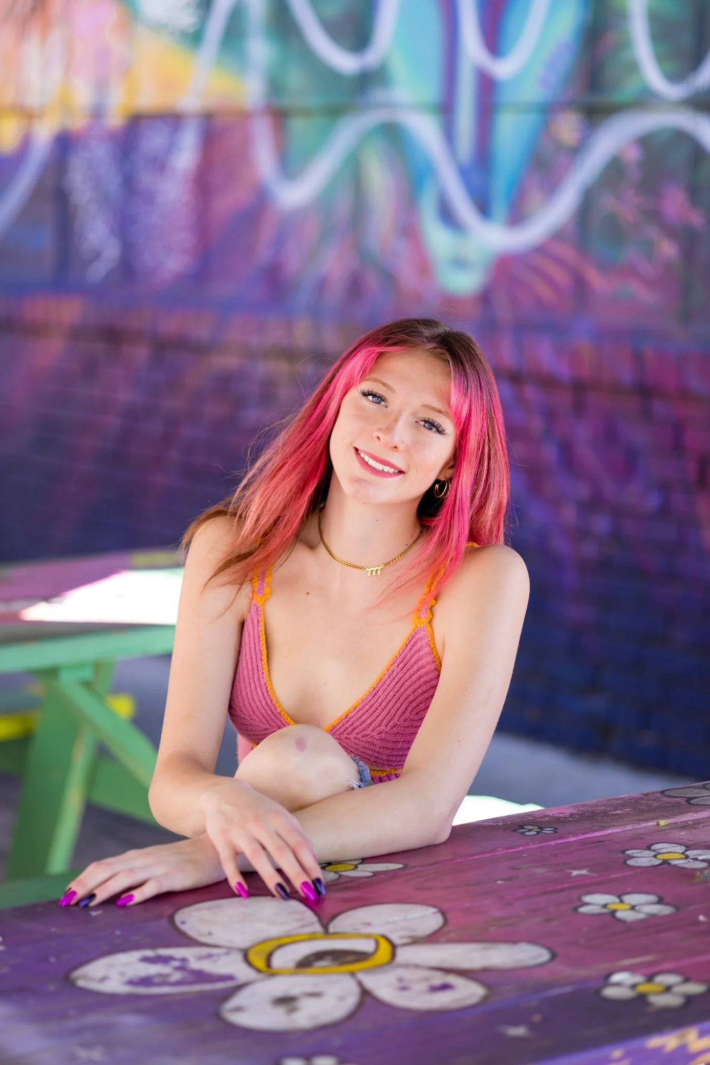 Young woman with pink hair smiling at camera, seated at a colorful, flower-decorated table against a vibrant graffiti-covered wall.