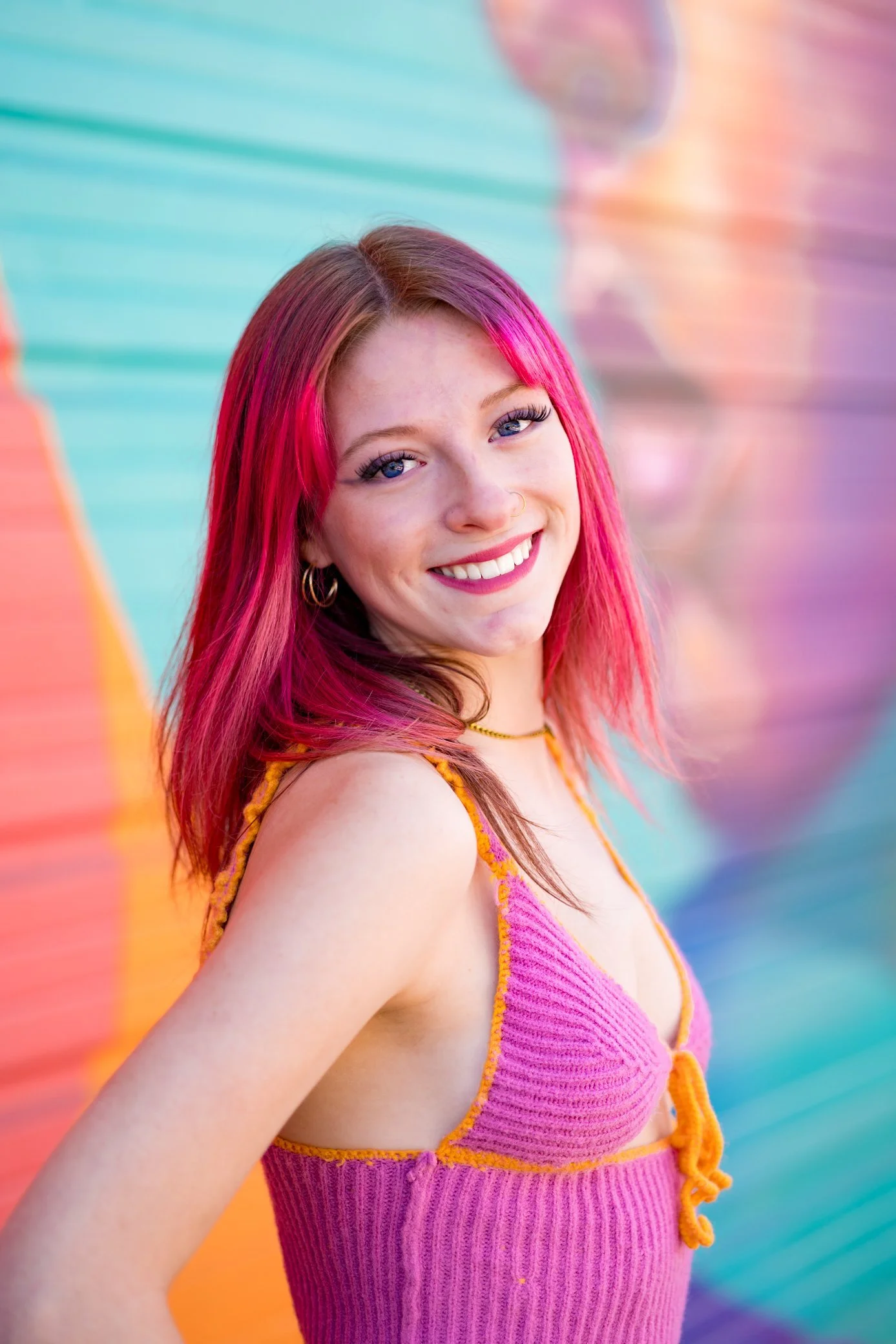 A young woman with pink hair, smiling, wearing a pink and orange sleeveless top, standing in front of a colorful rainbow mural.