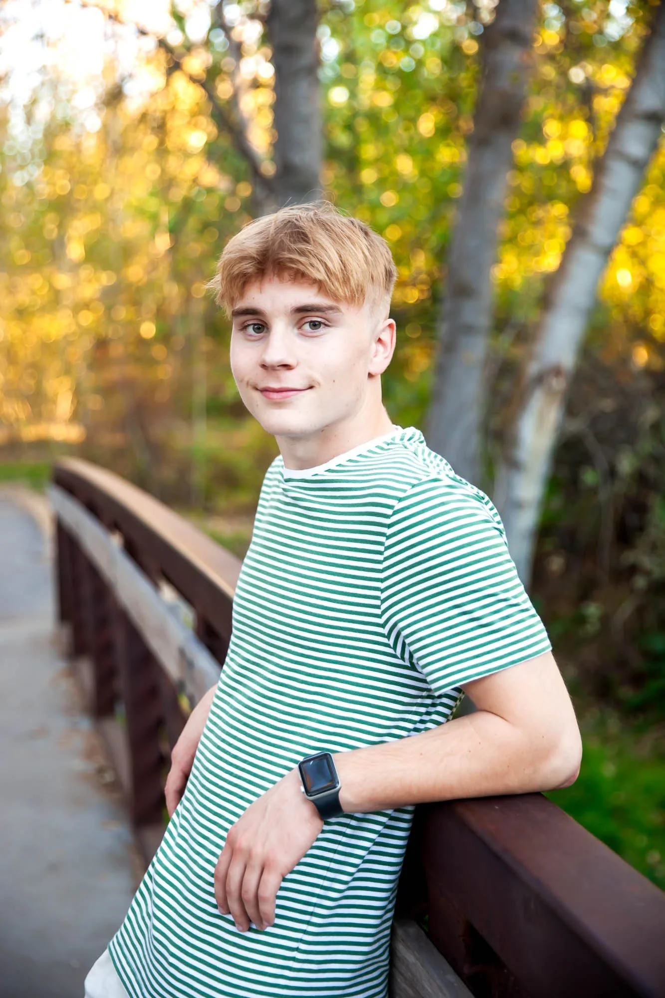 A young man with blond hair wearing a green and white striped t-shirt and black watch leaning against a wooden railing on a bridge, with trees and autumn foliage in the background.