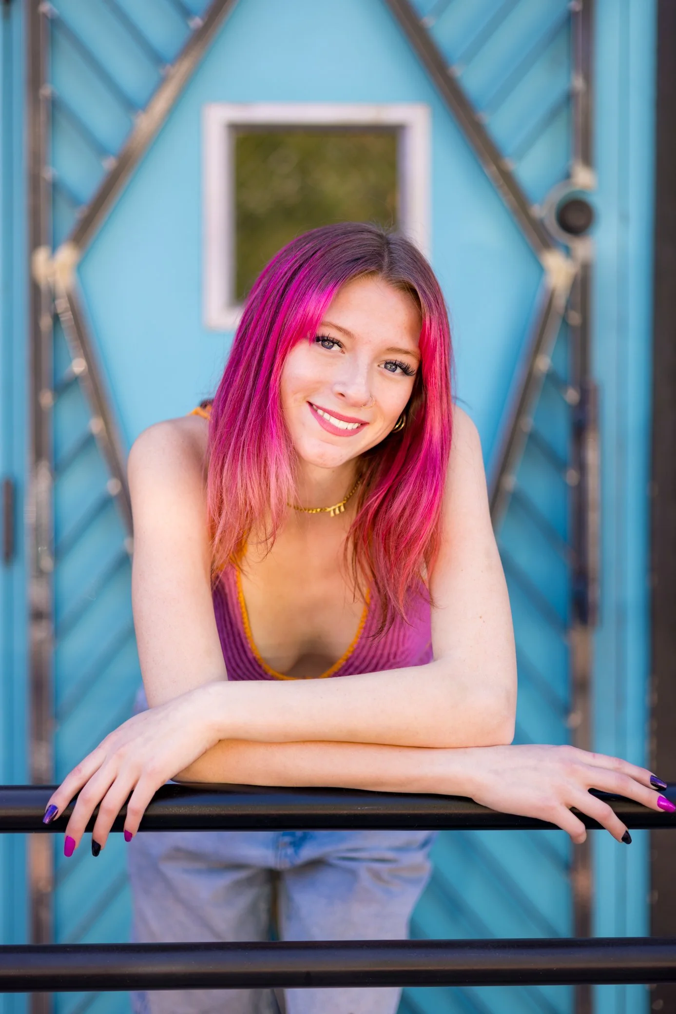 A young woman with pink hair smiling and leaning on a black railing in front of a colorful blue structure with a small window.
