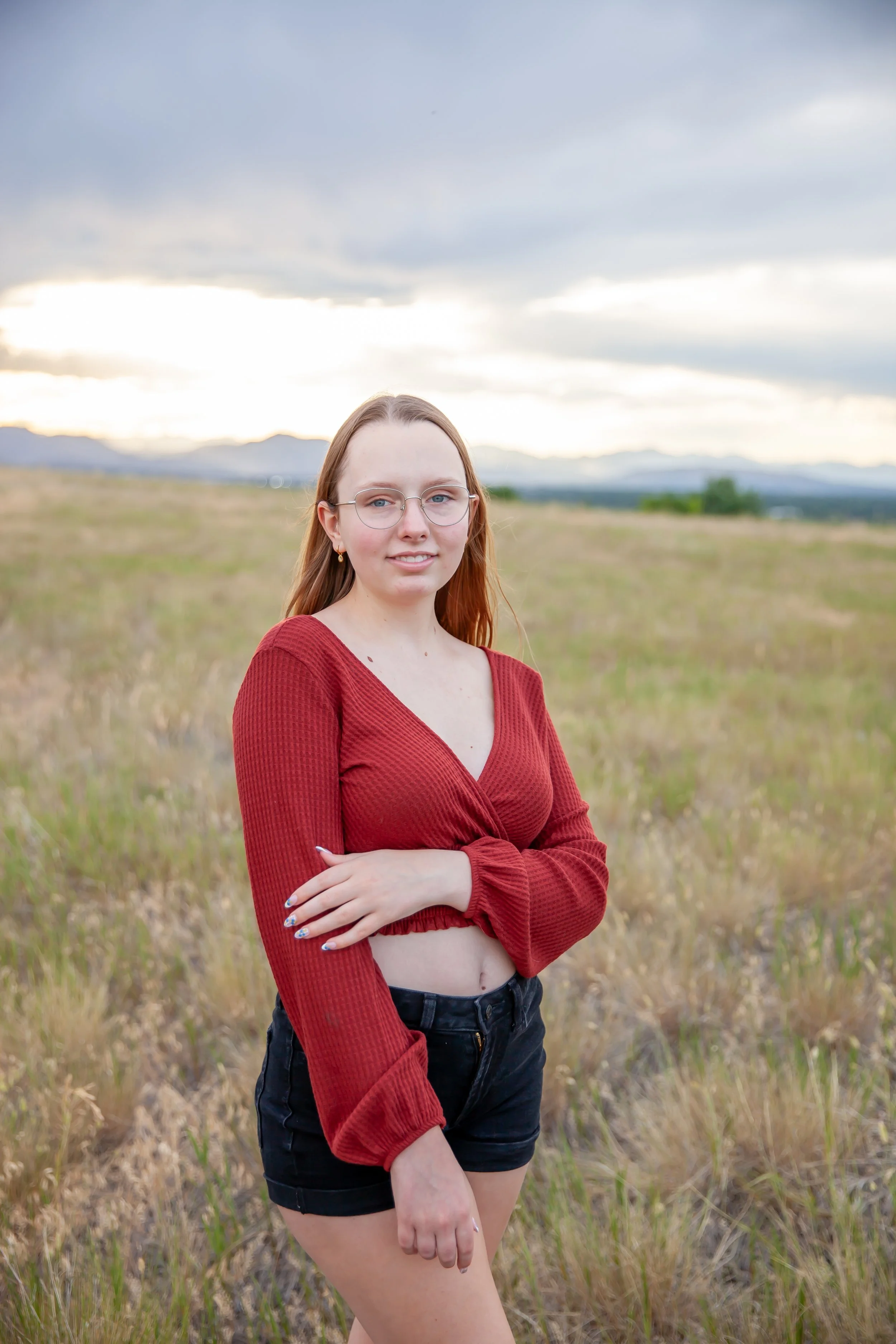 A young woman with long blonde hair, glasses, and light skin posing outdoors in a grassy field with mountains in the background and cloudy sky, wearing a red long-sleeve crop top and black shorts.