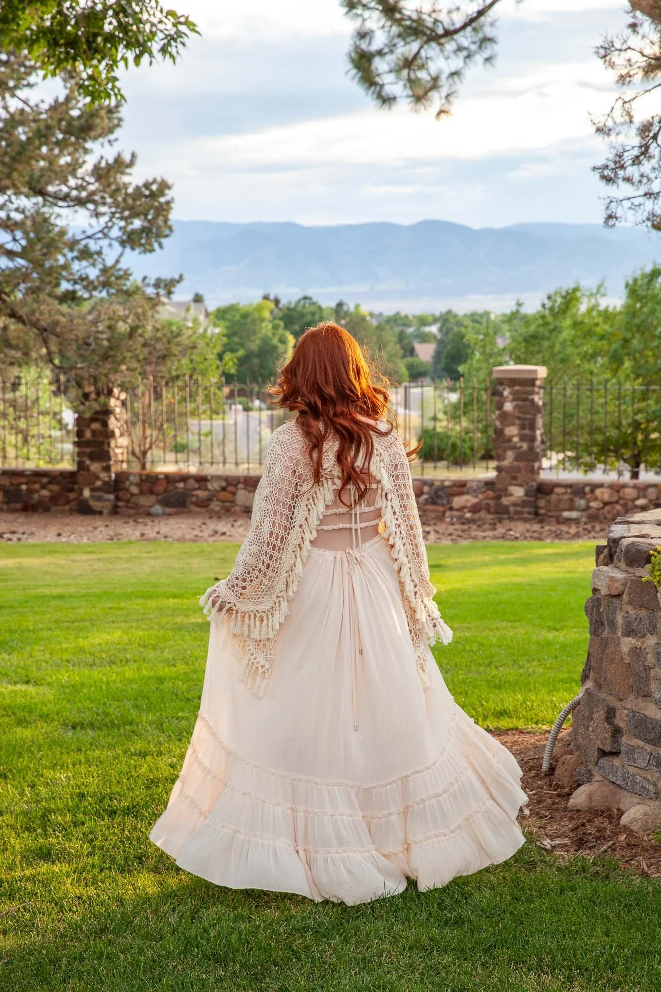 A woman with red hair in a white Victorian-style dress with a crocheted shawl, standing on a lush green lawn with a scenic view of mountains and trees in the background.