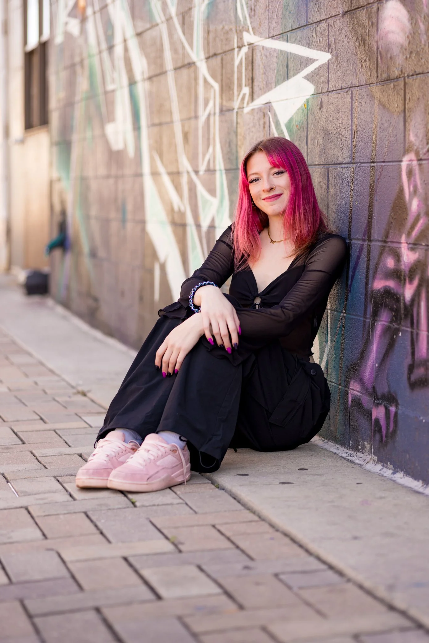 A young woman with pink hair sitting on a sidewalk against a graffiti-covered brick wall, smiling at the camera.