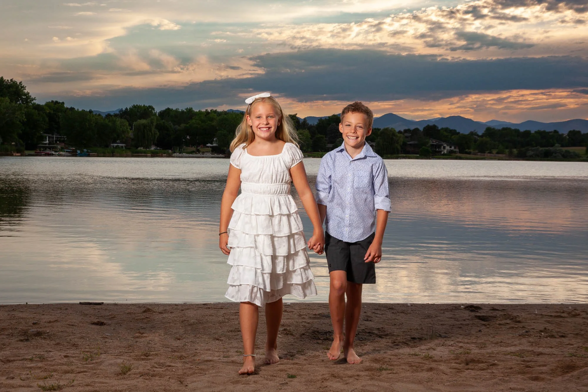 Two children, a girl in a white dress and a boy in a light blue shirt and dark shorts, walking hand-in-hand on a sandy beach near a lake with mountains and a cloudy sunset sky in the background.