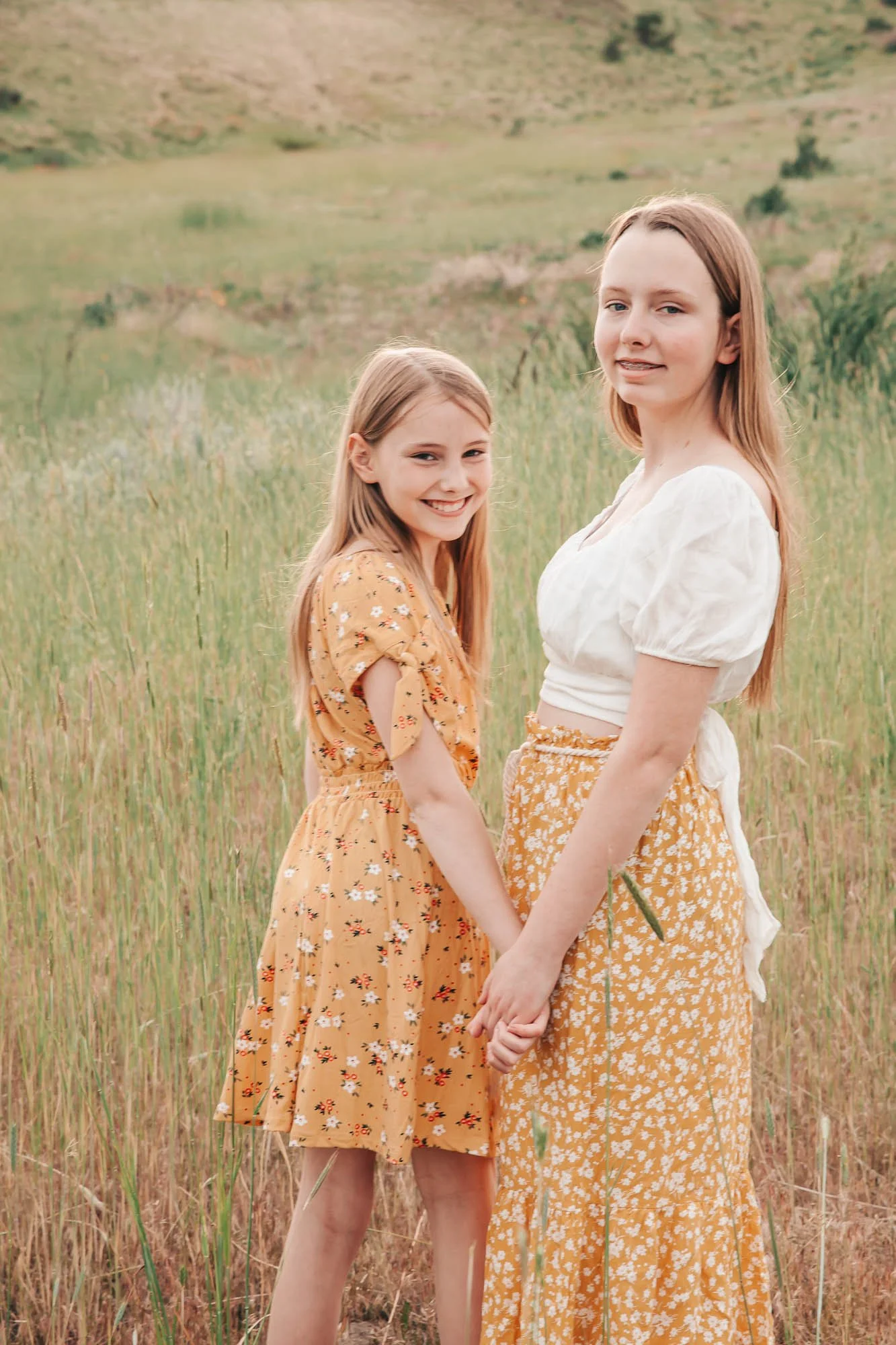 Two girls holding hands in a grassy field, wearing floral dresses.