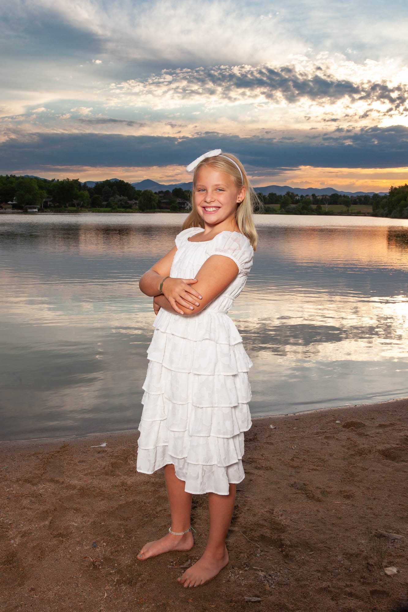 A smiling young girl with blonde hair wearing a white dress and a bow headband, standing barefoot on a sandy beach by a lake at sunset with a partly cloudy sky and distant mountains.