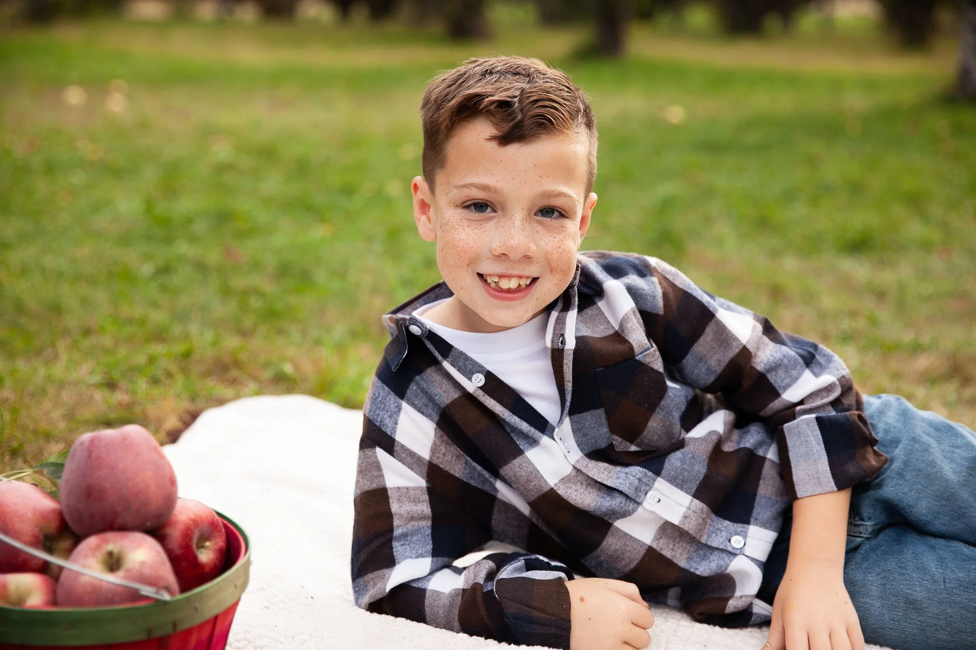 A young boy with brown hair and freckles lying on a white blanket outdoors, smiling at the camera with a bowl of apples nearby.