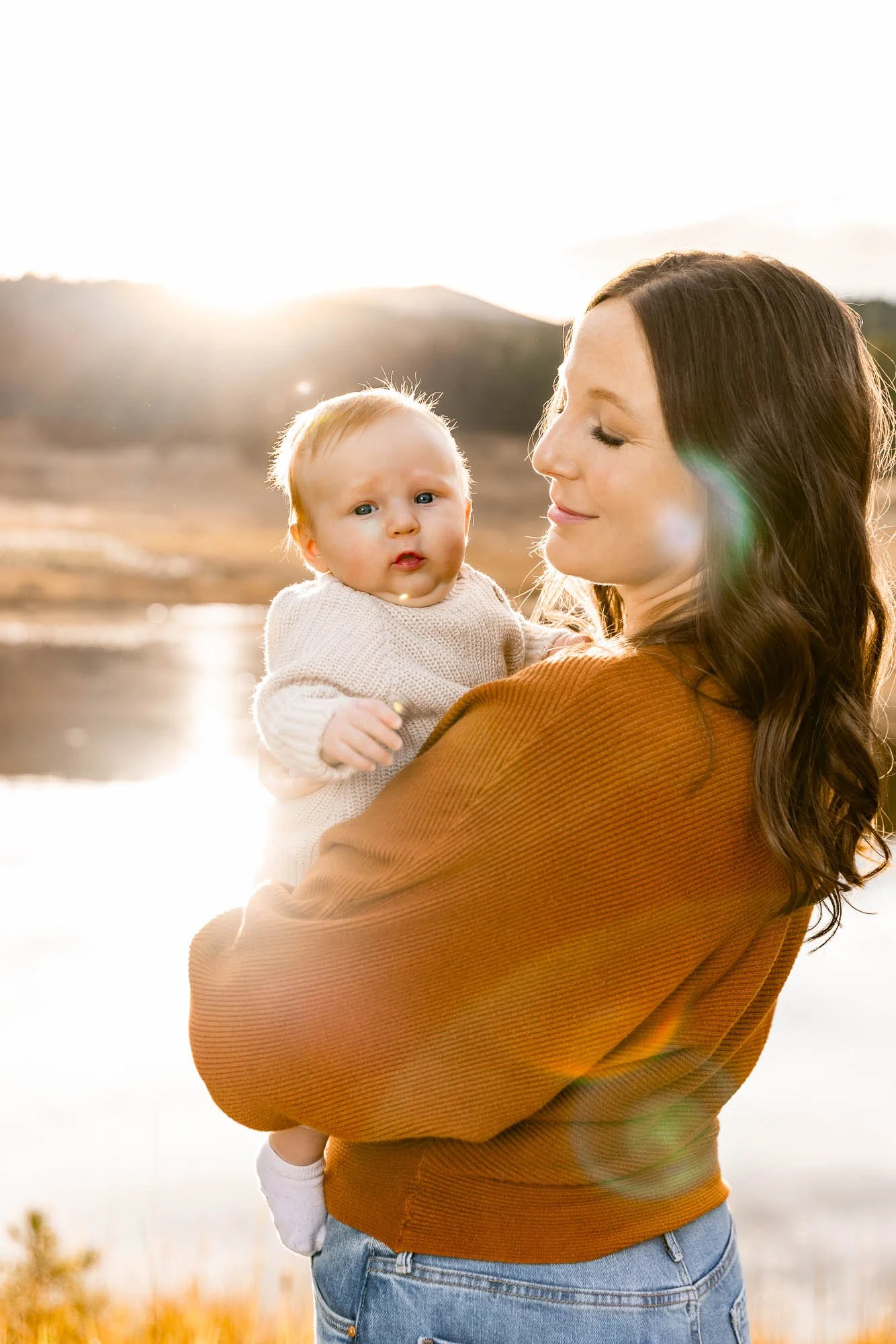 A woman holding a child outdoors during sunset with hills in the background.