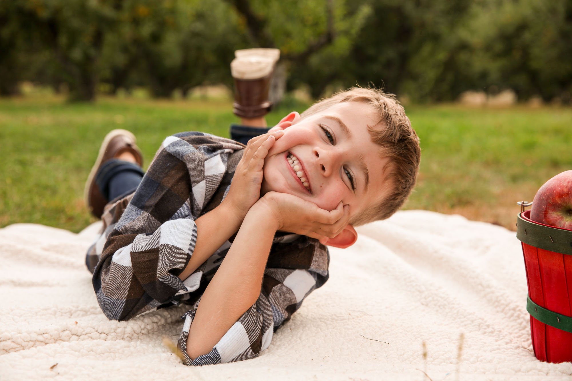 A young boy with curly hair lies on his stomach on a cream-colored blanket outdoors, smiling and resting his head on his hands. He is wearing a checkered shirt and jeans. There is a red picnic basket with green straps and a metal latch on the right s
