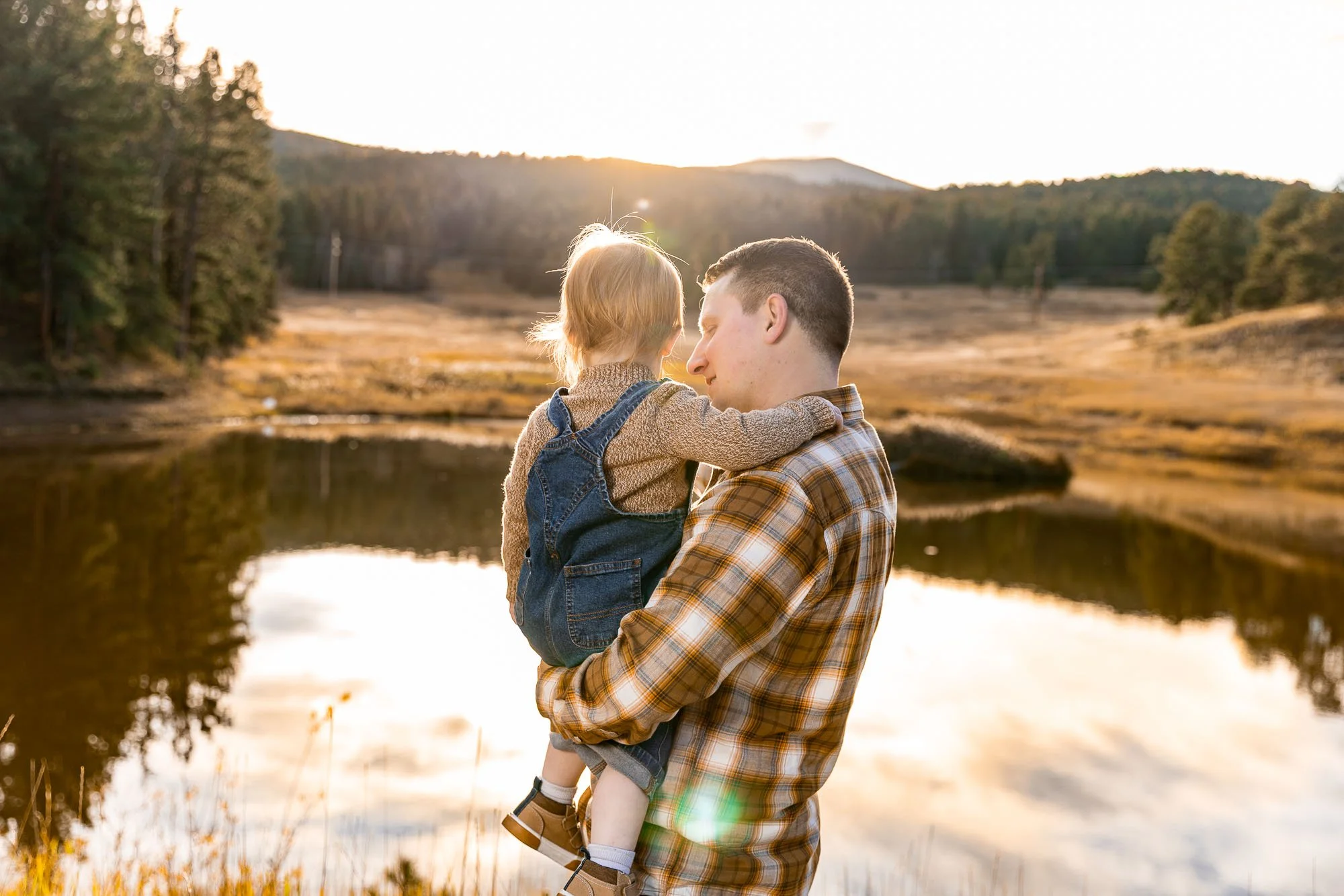 A man holding a young girl near a lake at sunset, surrounded by trees and mountains in the background.