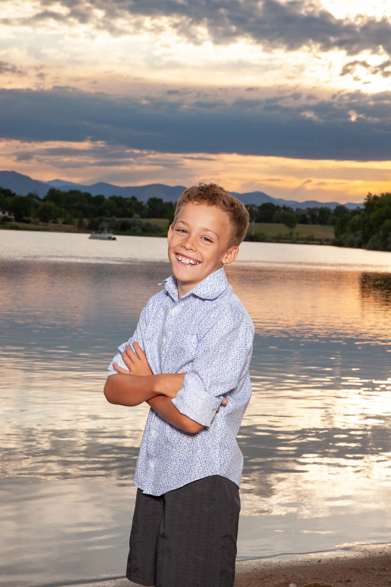 A young boy with a big smile, arms crossed, standing on the beach at sunset with a lake and mountains in the background.