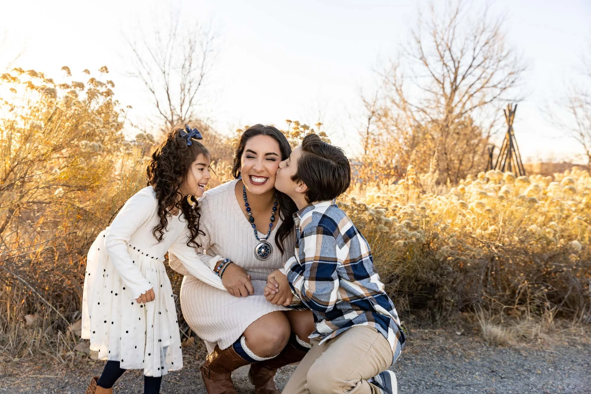 A woman with two children smiling and laughing outdoors in a fall landscape, with trees and bushes with golden leaves.