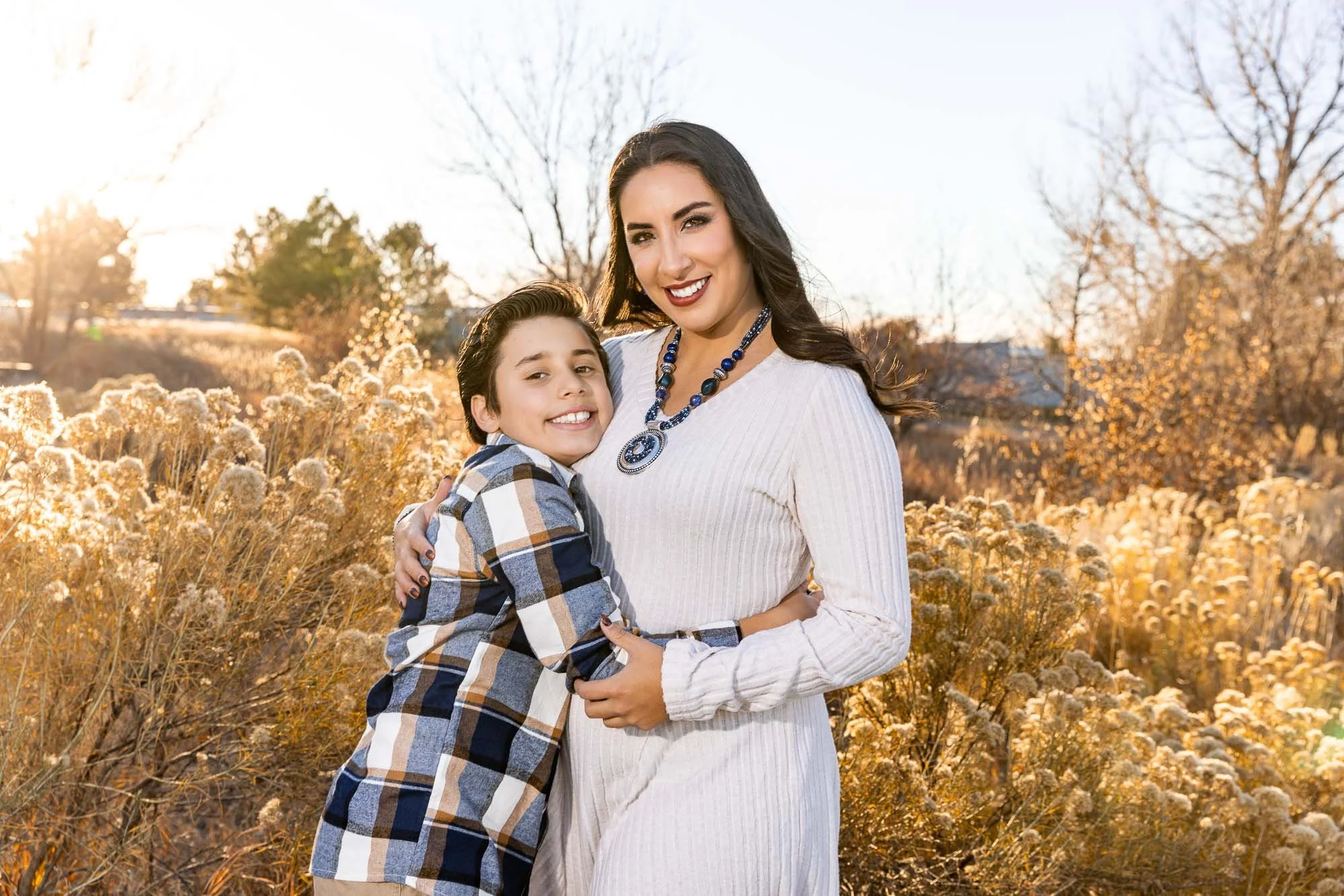 A smiling woman with long dark hair wearing a white dress and a blue necklace hugging a young boy with dark hair, wearing a plaid shirt, in a field of dried flowers during golden hour with trees in the background.