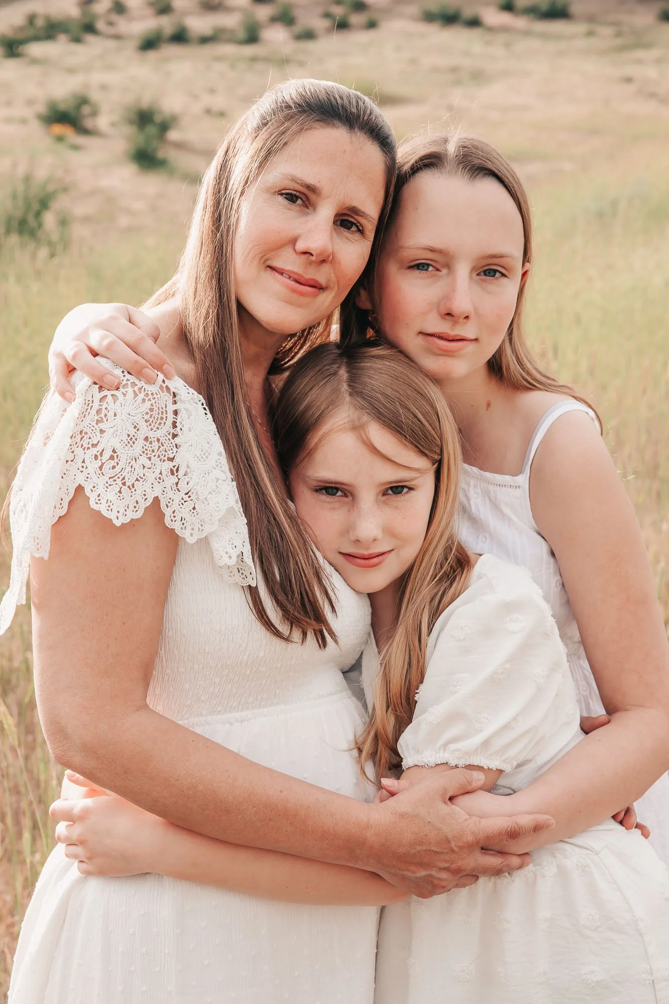 Mother and two daughters in white dresses embracing outdoors in a field.