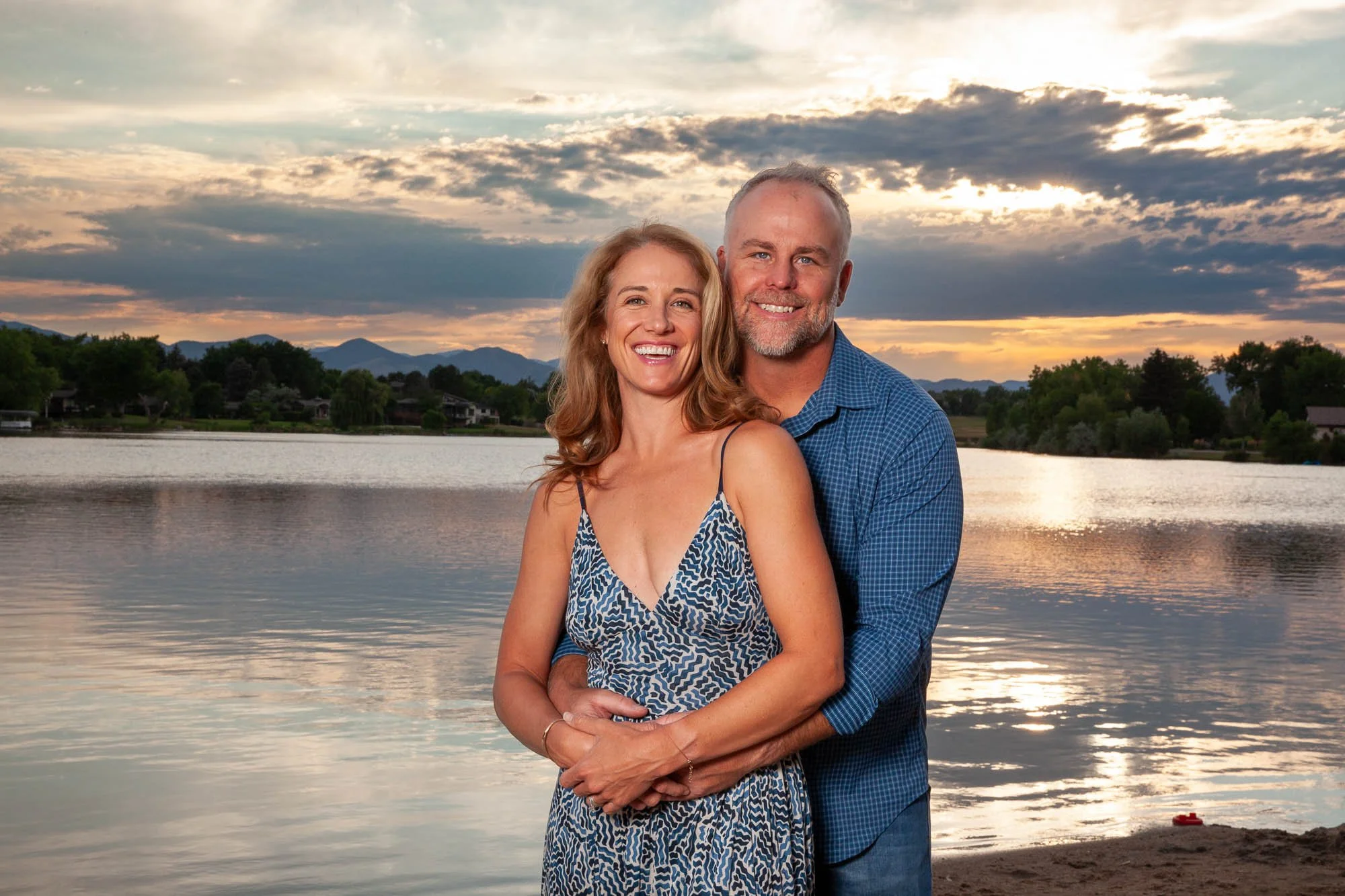 A smiling couple stands arm in arm by a lakeshore at sunset, with mountains and trees in the background.