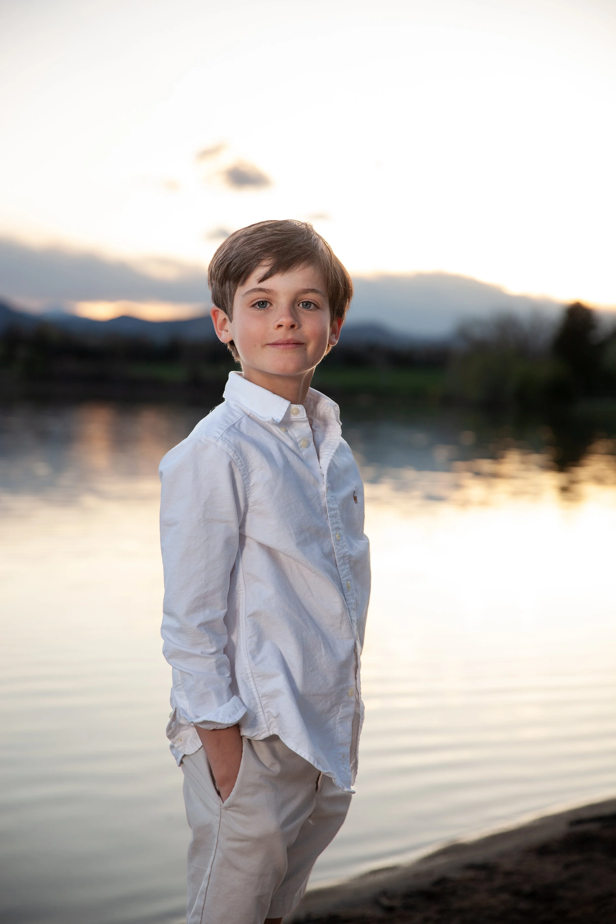 A young boy standing by a lake during sunset, wearing a white shirt and light-colored pants, with hands in pockets. The background features a calm lake and distant hills under a cloudy sky.