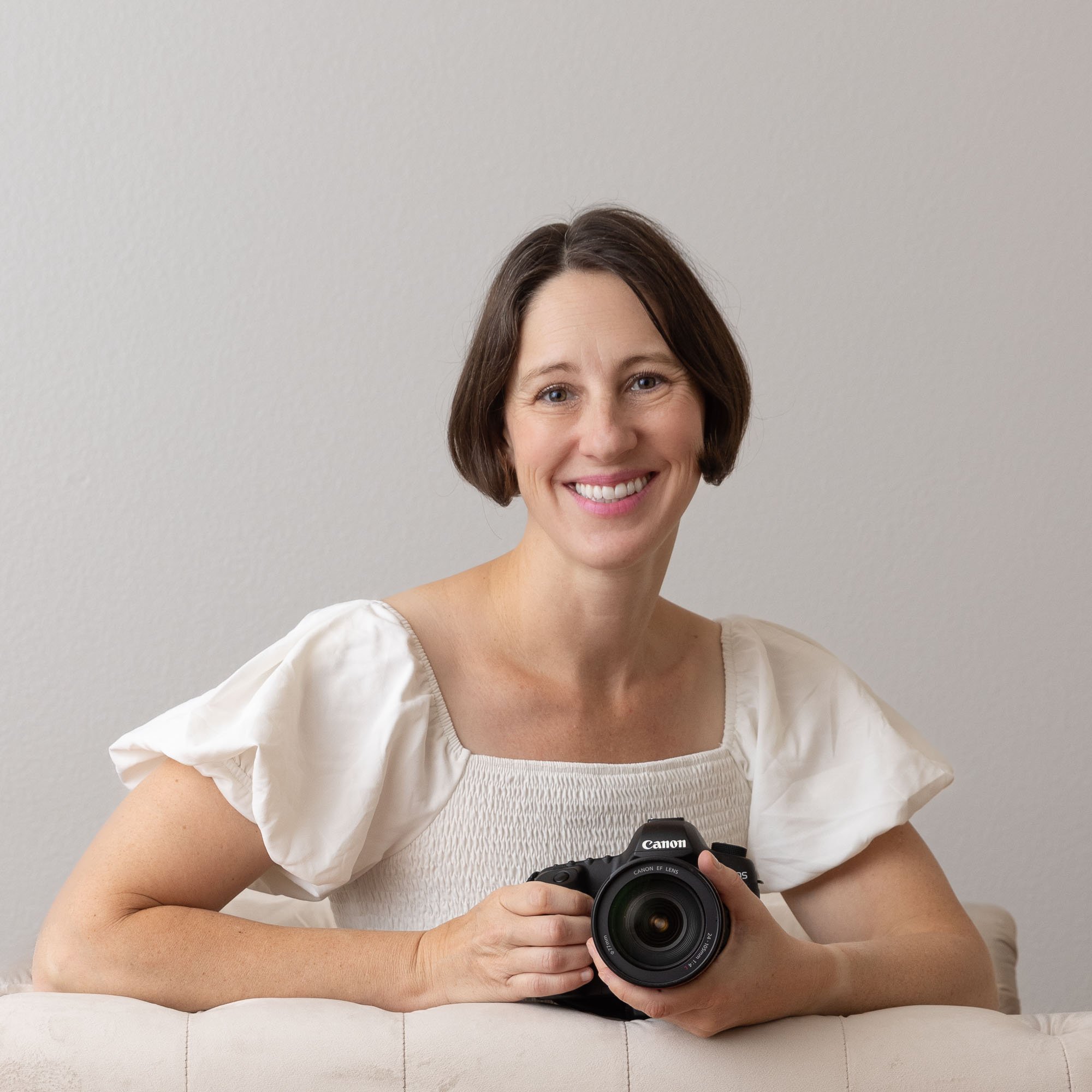 A woman with short brown hair, wearing a white blouse, smiling and holding a Canon camera.