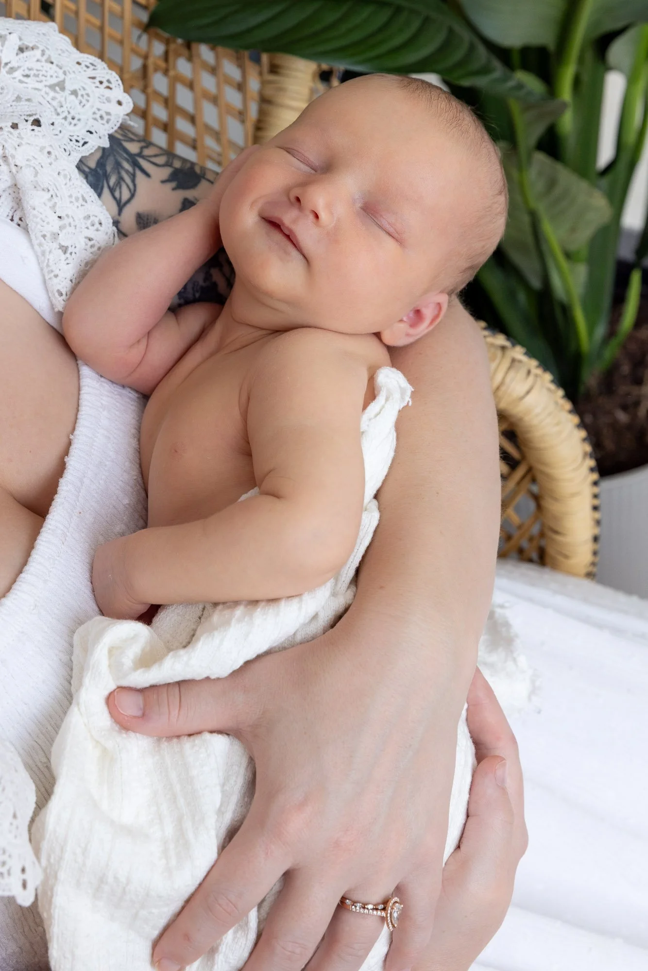 A sleeping baby with closed eyes, resting on an adult's arm. The baby has light skin, faintly visible eyebrows, and a small amount of hair. The adult's hand, wearing a gold ring with small diamonds, gently supports the baby's body. A white cloth or b