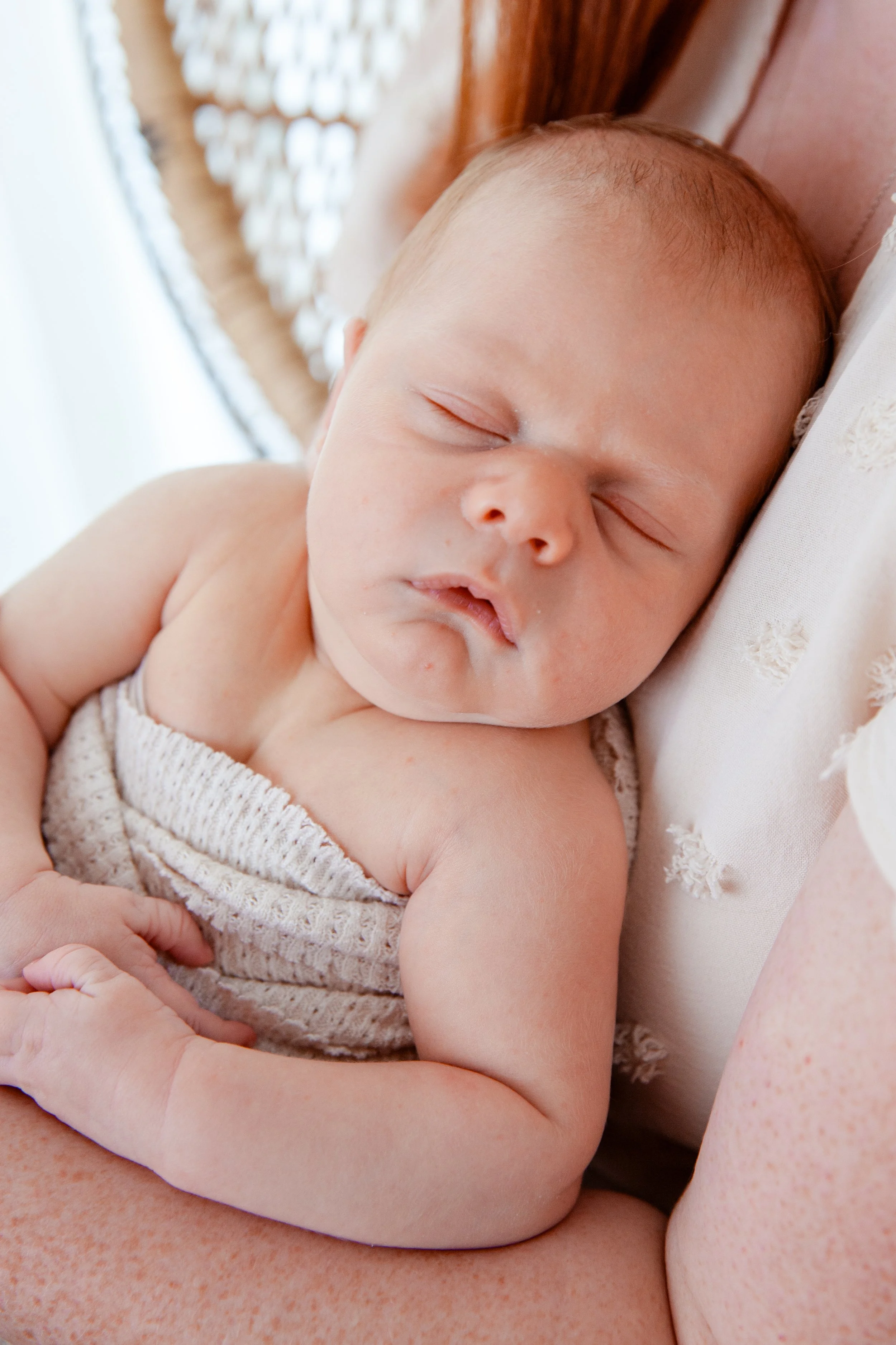 Close-up photo of a sleeping baby with a peaceful expression, lying in someone's arms, wearing a sleeveless knitted garment.