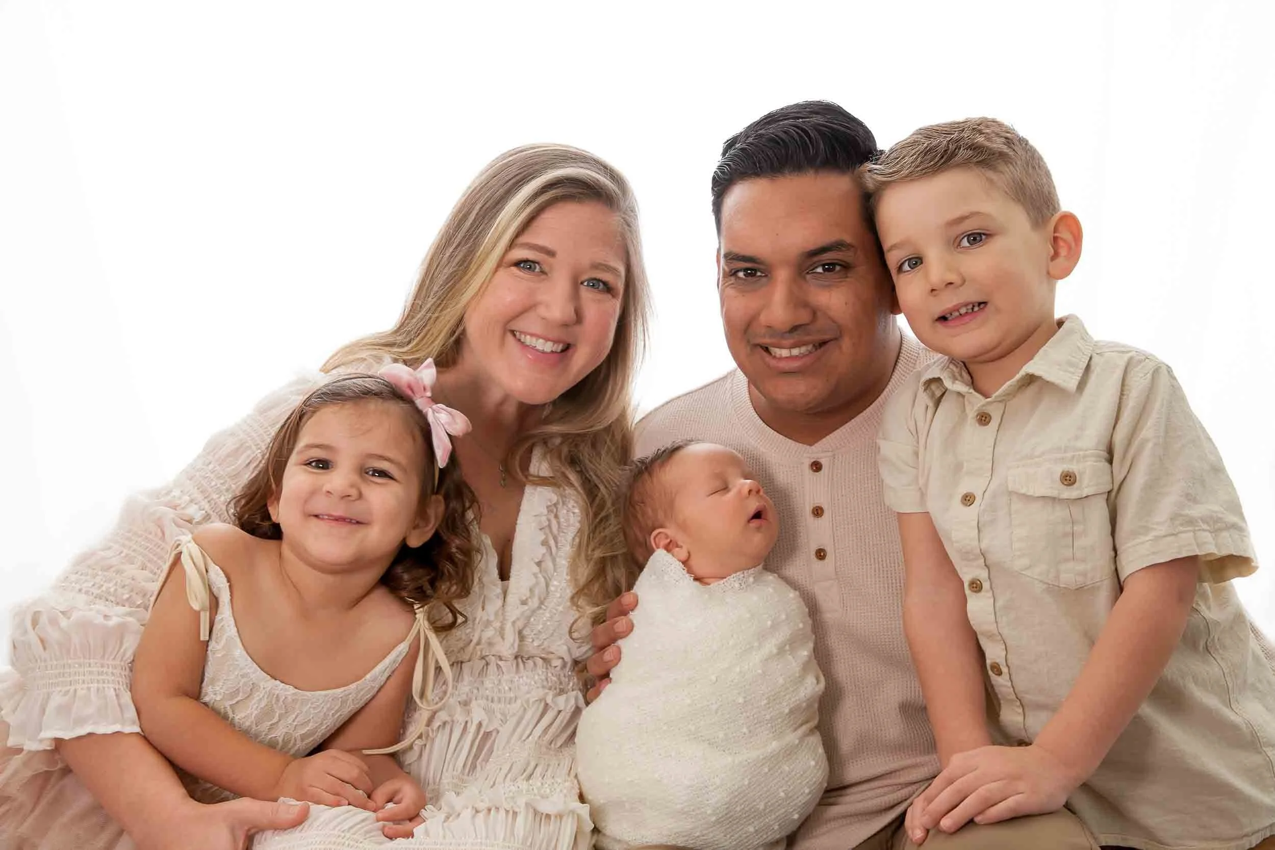 A family of five with three children, including one infant, is smiling and posing together. The adults are holding the baby, and everyone is wearing light-colored, casual clothing, standing against a plain white background.