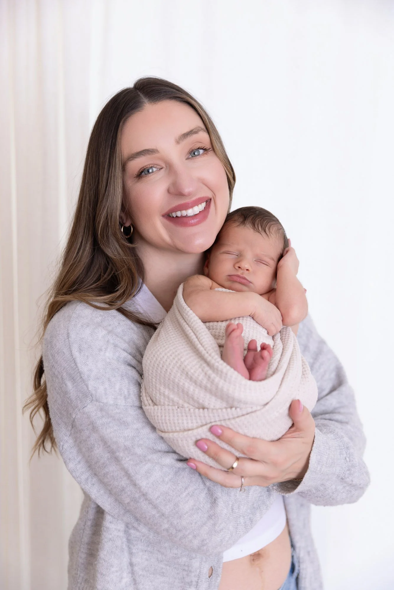 A woman with long brown hair, blue eyes, and a big smile holding a sleeping baby wrapped in a beige blanket, against a plain white background.