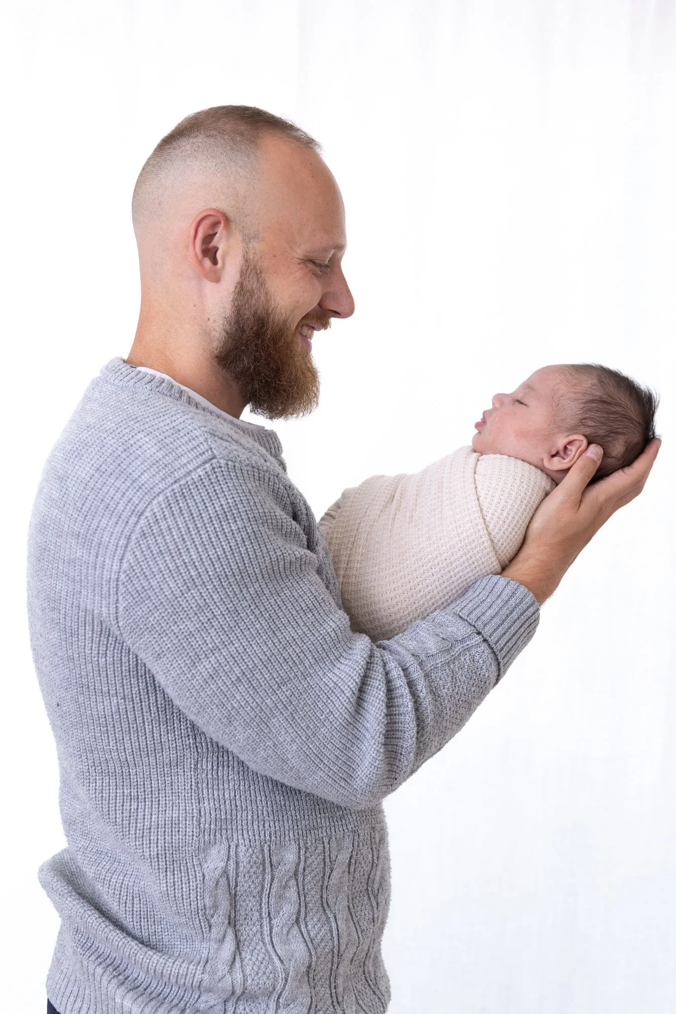 A man with a beard and short hair smiling while holding a baby wrapped in a beige blanket, against a plain white background.