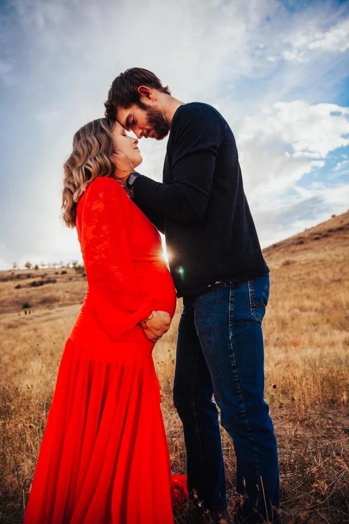 Pregnant woman in red dress embracing man in outdoor setting with hills and sky.