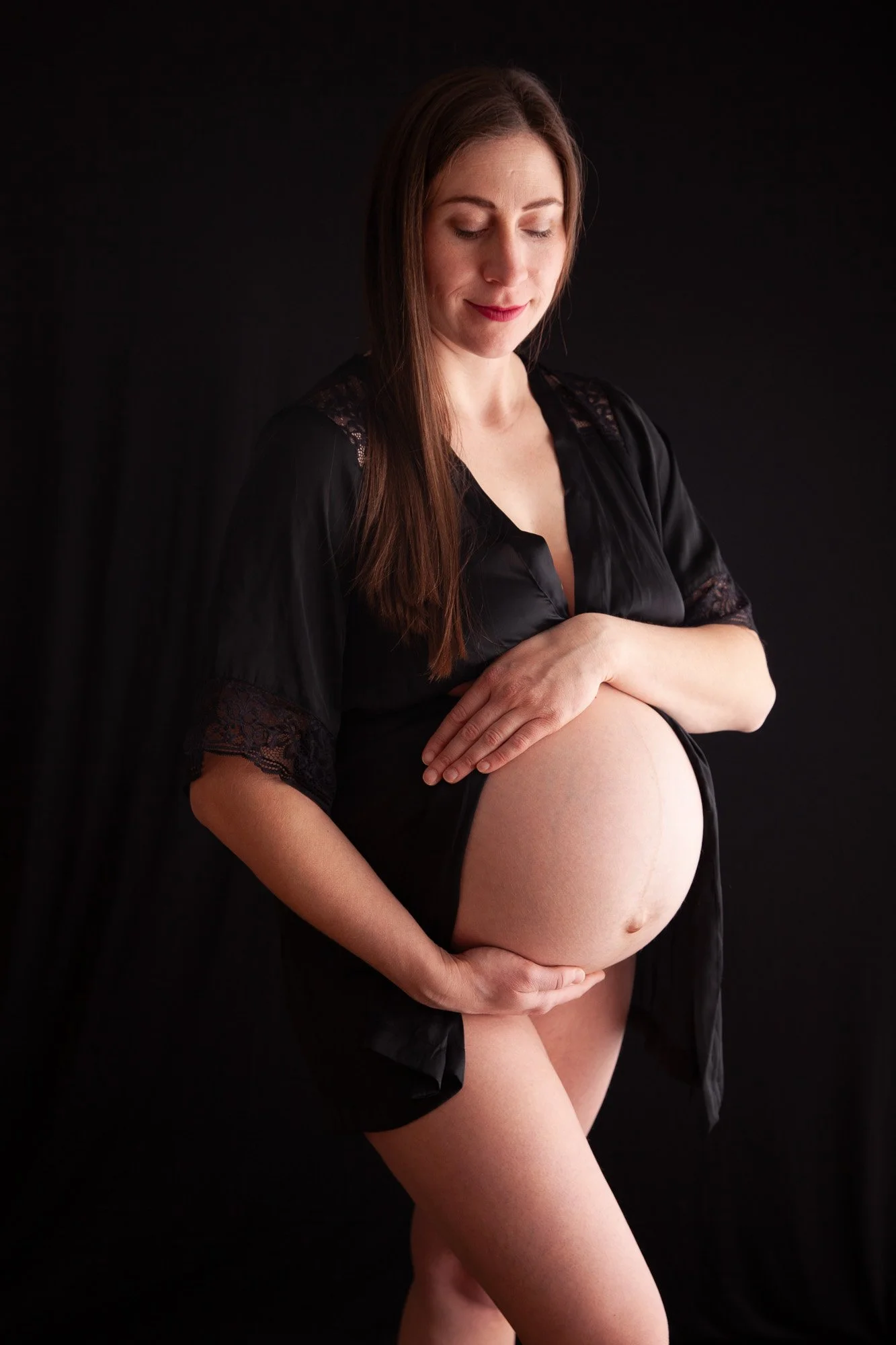A pregnant woman in a black robe, gently holding her large belly, standing against a dark background with her eyes closed and a peaceful expression.