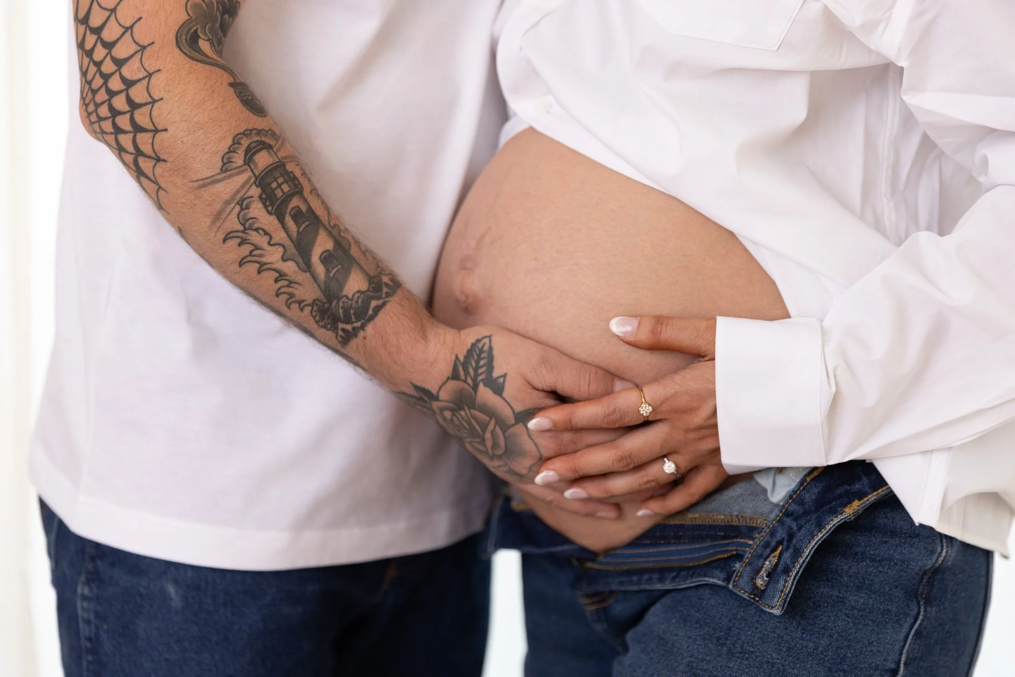 Close-up of a pregnant woman and her partner holding her belly, showing their hands and part of their torsos, with the woman wearing rings and the man having tattoos on his arm.