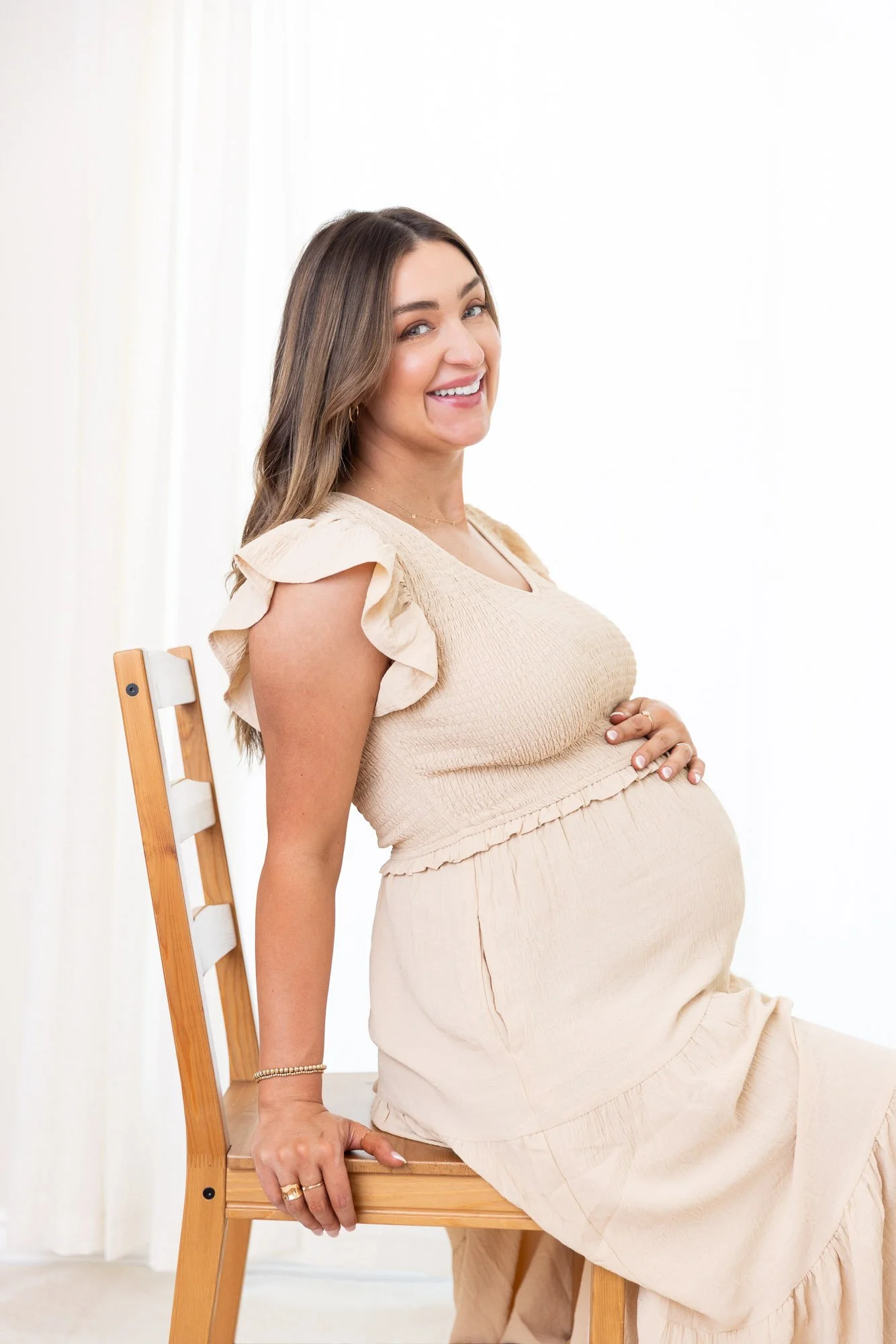 A pregnant woman sitting on a wooden chair, smiling and looking at the camera, wearing a light beige dress with ruffled sleeves in a bright, minimalistic room.