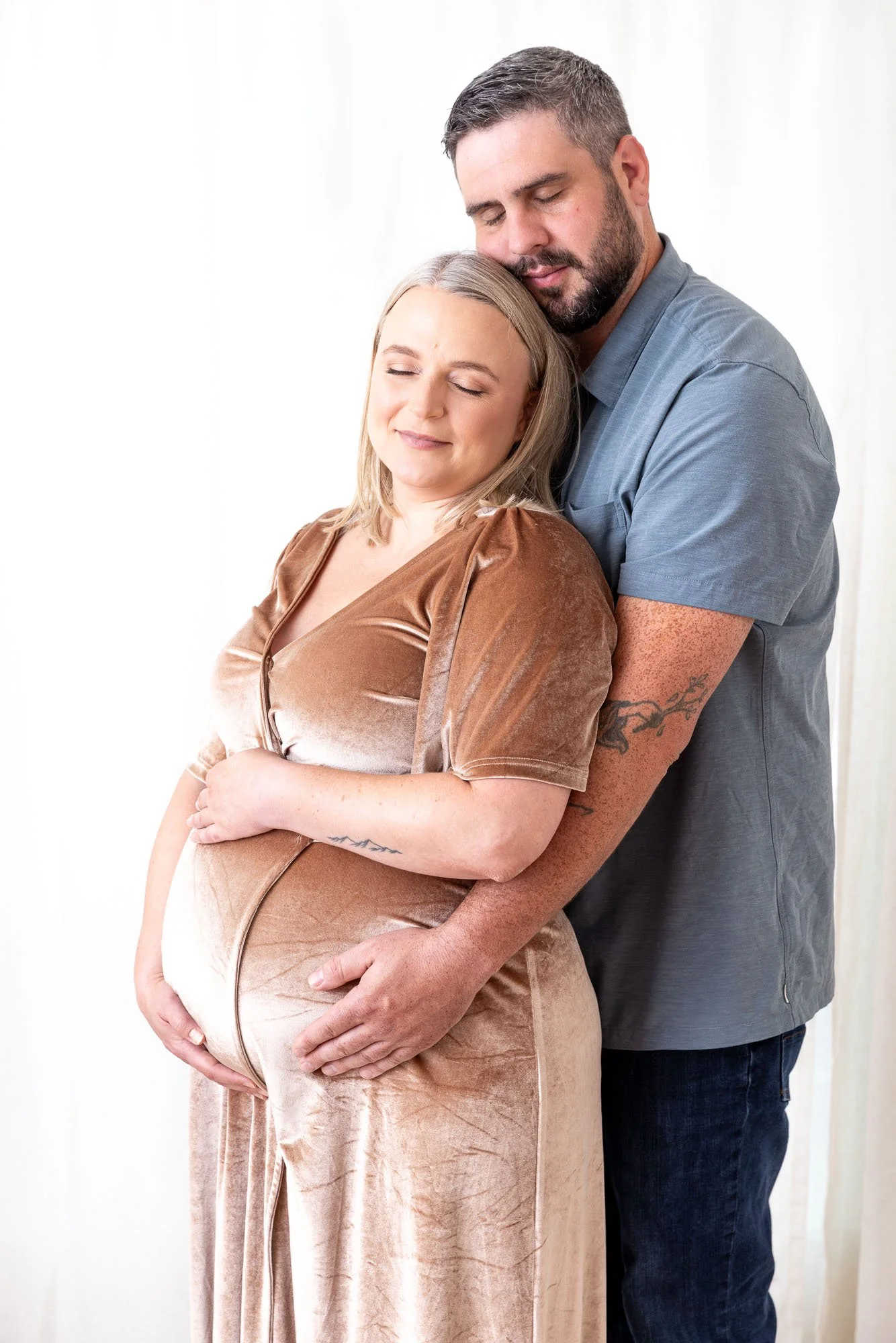 A smiling pregnant woman with blonde hair, wearing a brown velvet dress, is embraced by a man with a beard and short hair, wearing a gray shirt. The man is gently holding her belly. They are standing in front of a white background.