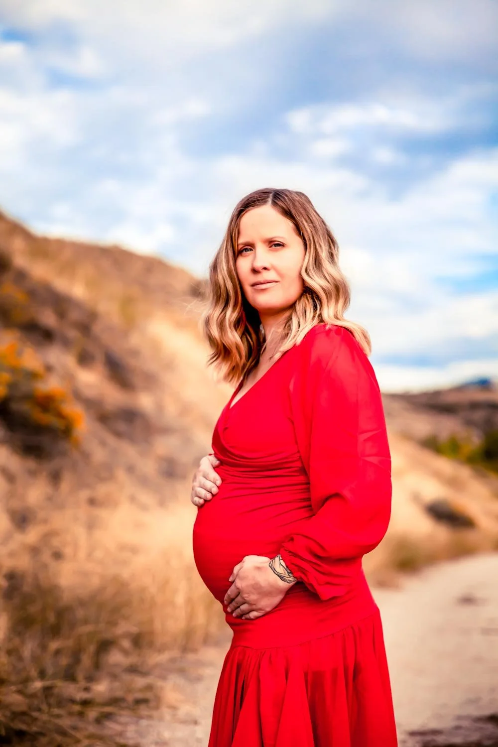 Pregnant woman in a red dress standing outdoors on a dirt path with hills and a cloudy sky in the background.