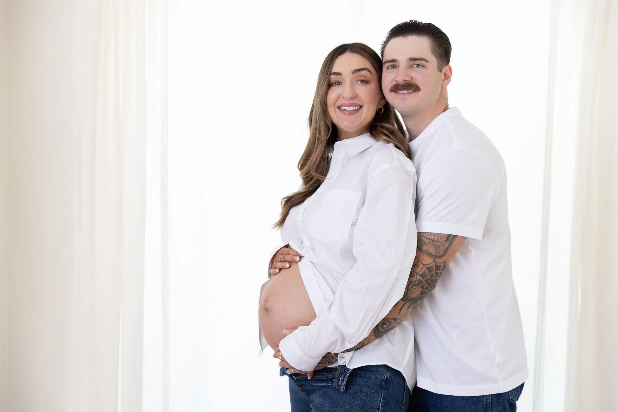 A smiling pregnant woman and man standing together indoors, with the woman showcasing her pregnant belly, both wearing white shirts and jeans.