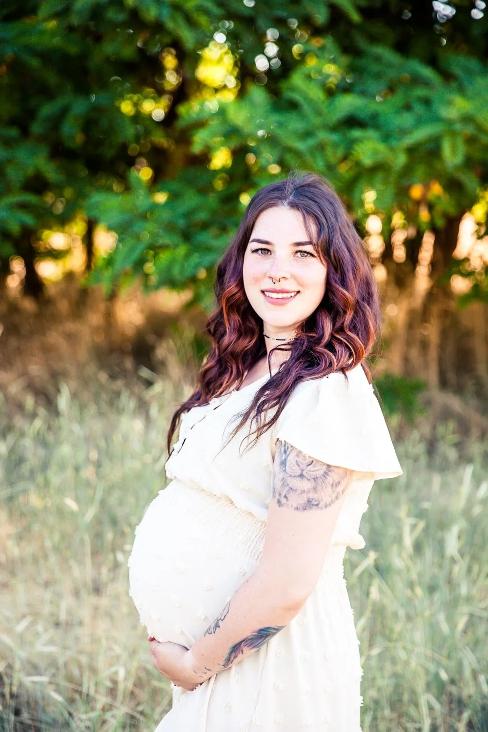 A pregnant woman with burgundy and black hair, tattoos on her arm, smiling outdoors during sunset, standing in grassy field with green trees in background.
