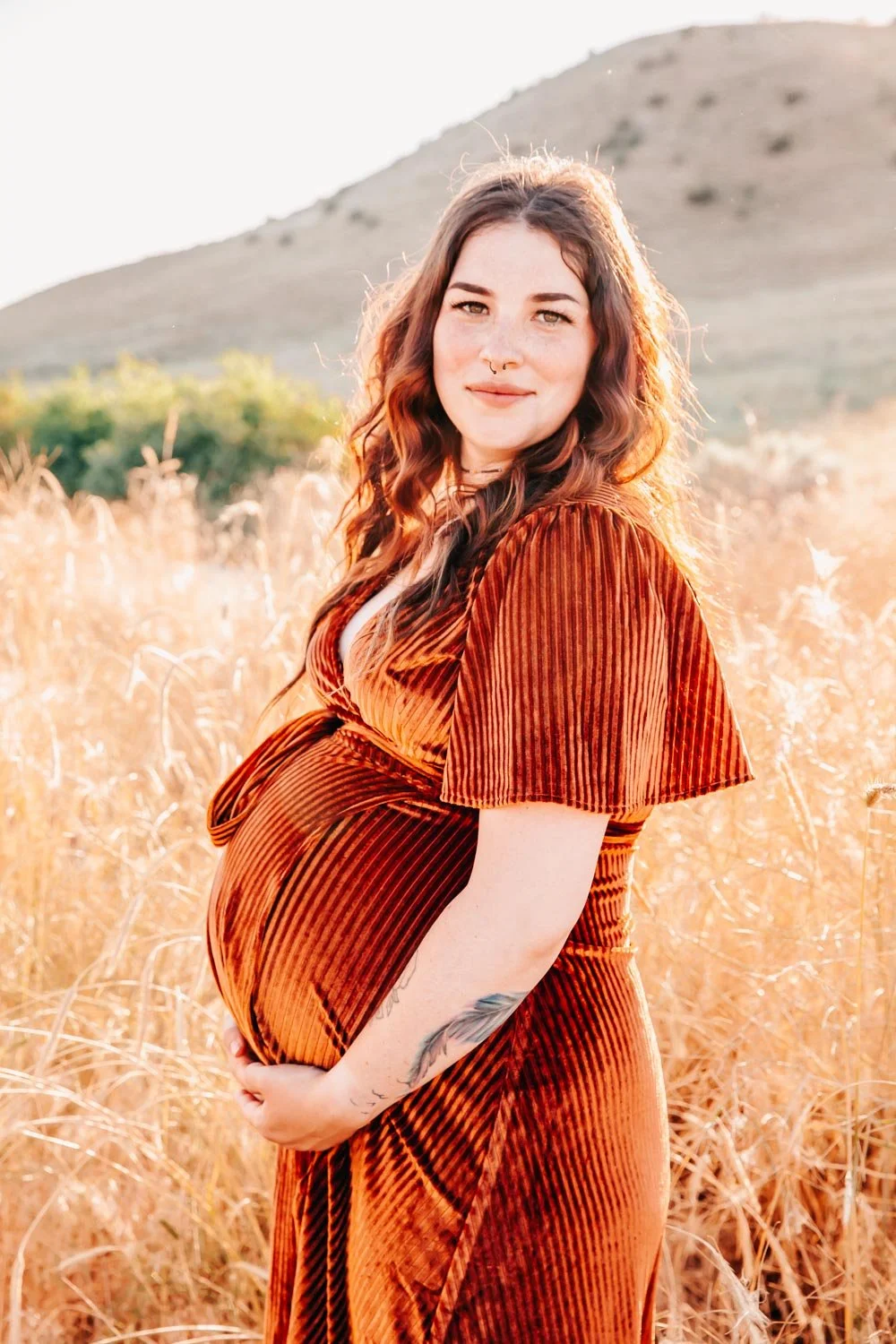 Pregnant woman in an orange dress standing in a field with hills in the background.