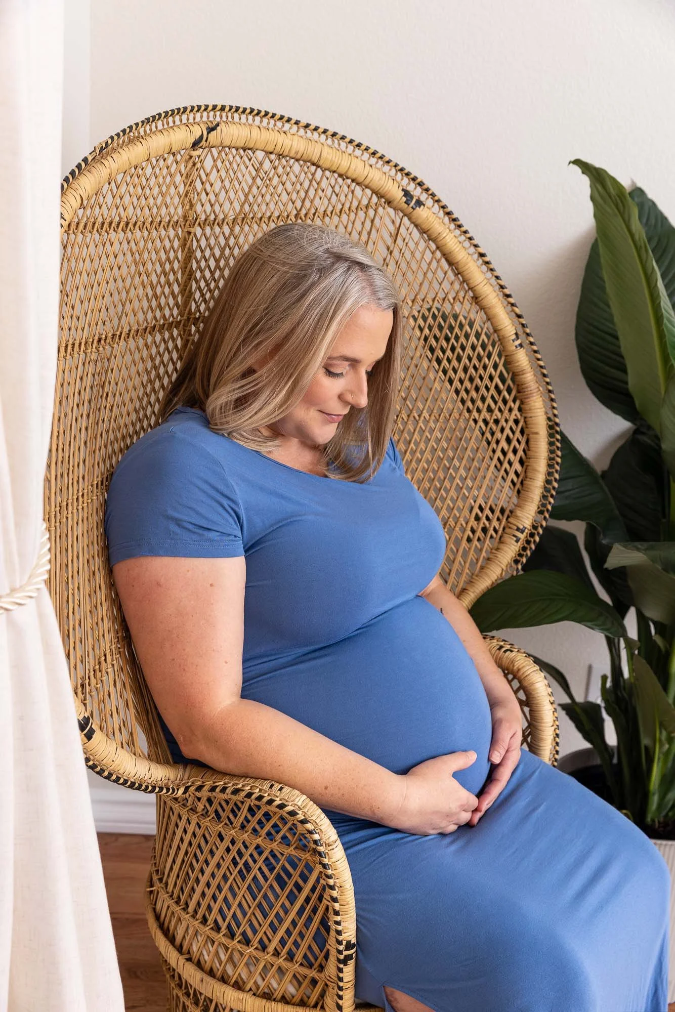 Pregnant woman sitting in a wicker chair with hands on her belly, wearing a blue dress, near a leafy plant and a white wall.