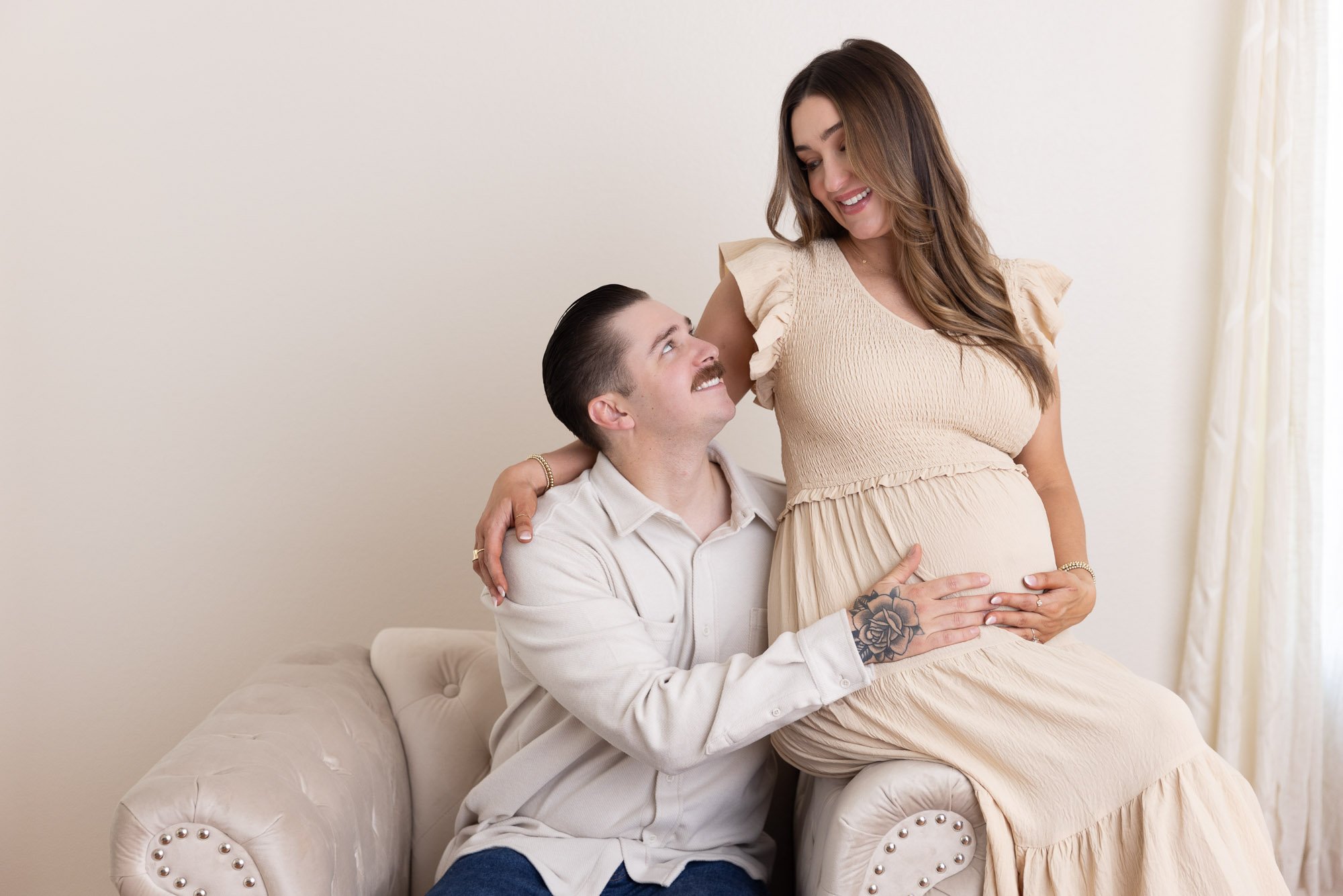 A man with a tattoo on his hand and a woman with long hair sitting on a beige couch, the man is touching the woman's pregnant belly while they smile at each other.