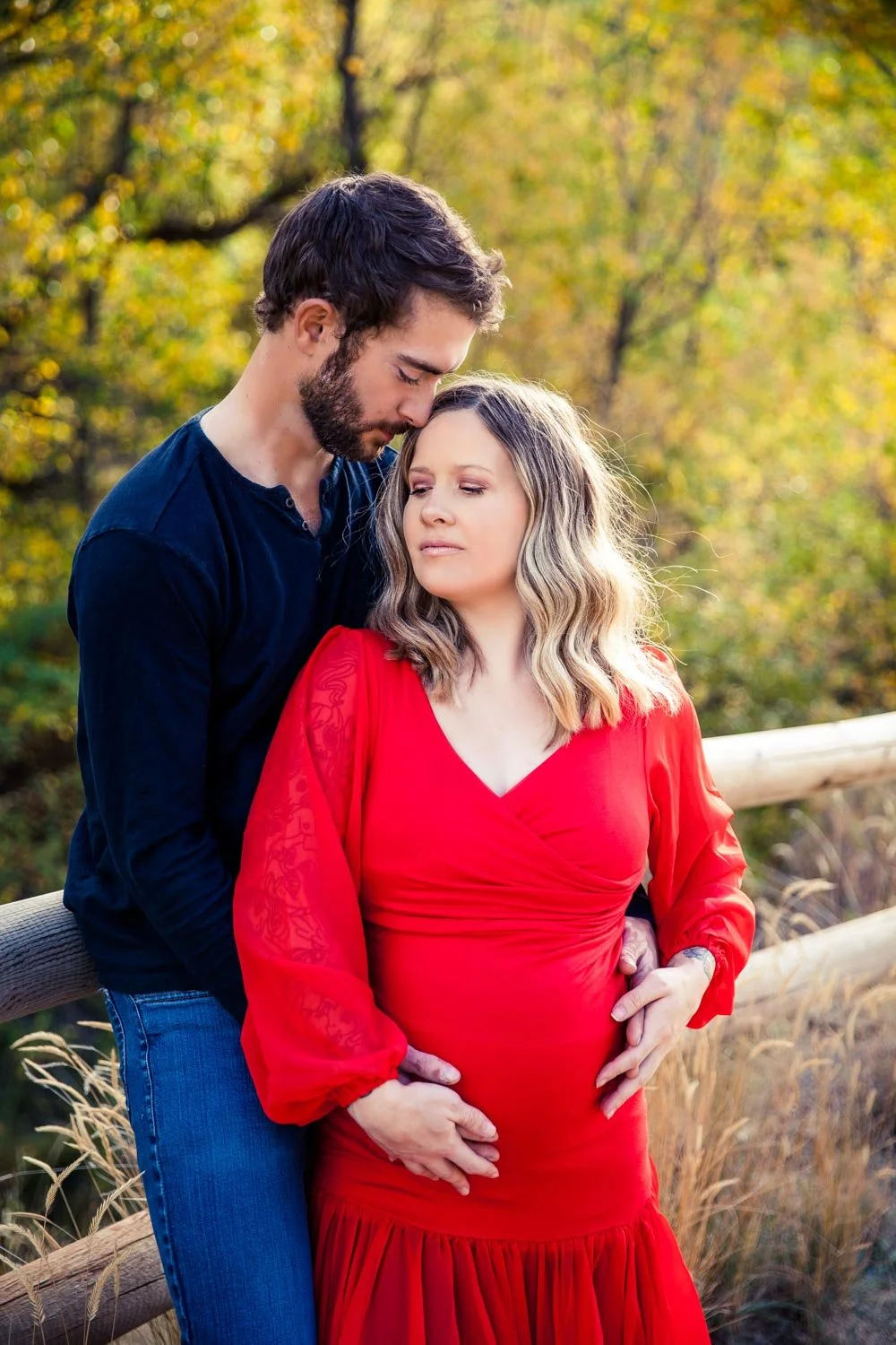 A man embraces a pregnant woman in a red dress in an outdoor setting with a wooden fence and trees in the background.