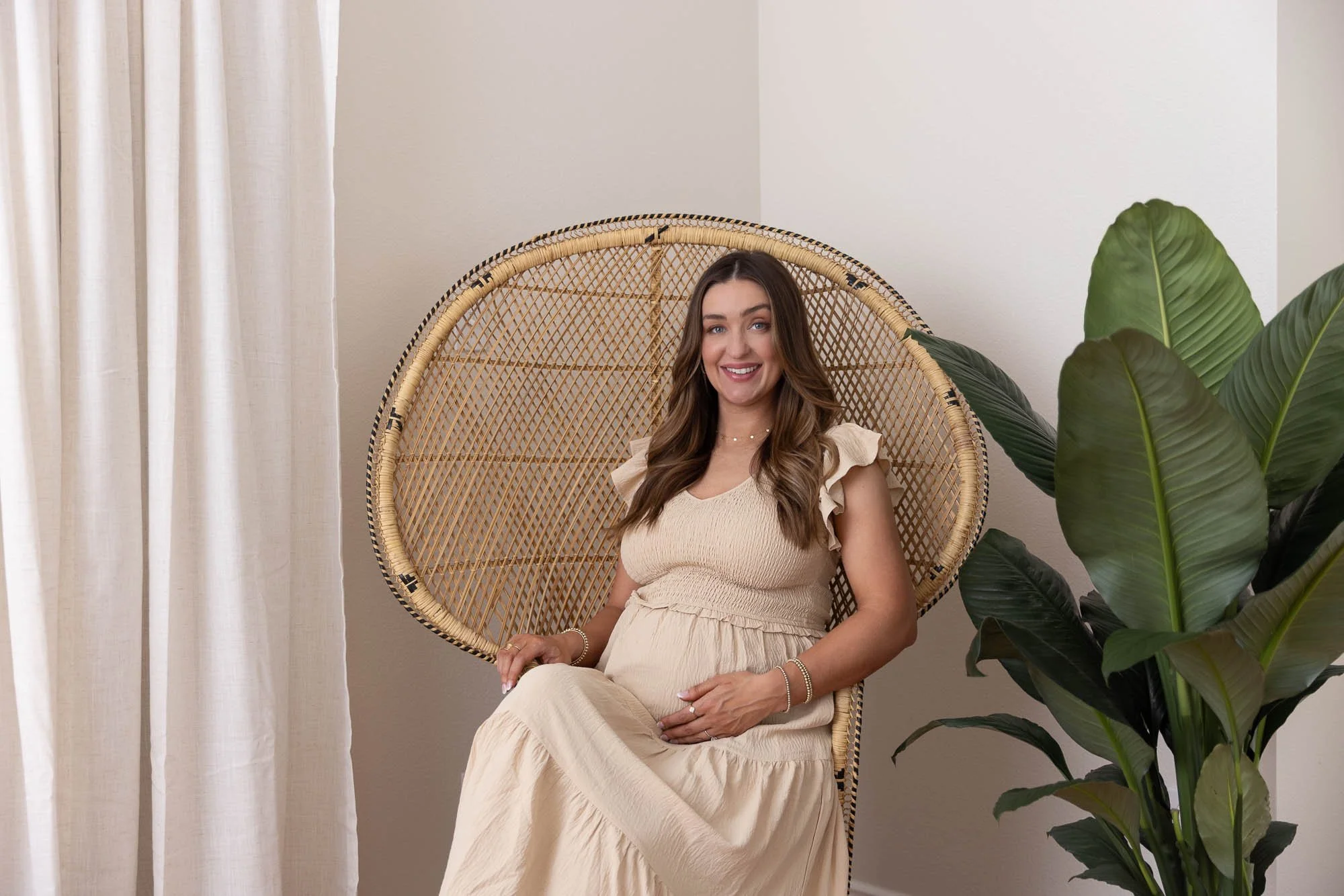 A pregnant woman with long brown hair sitting in a wicker peacock chair in a bright room with white walls and curtains, next to a large green houseplant, smiling and holding her baby bump.
