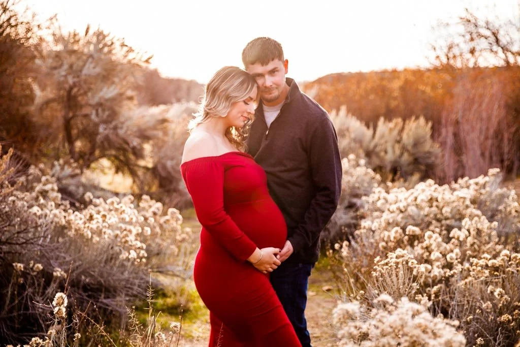 Pregnant woman in red dress and man standing together in a field with dry plants at sunset.