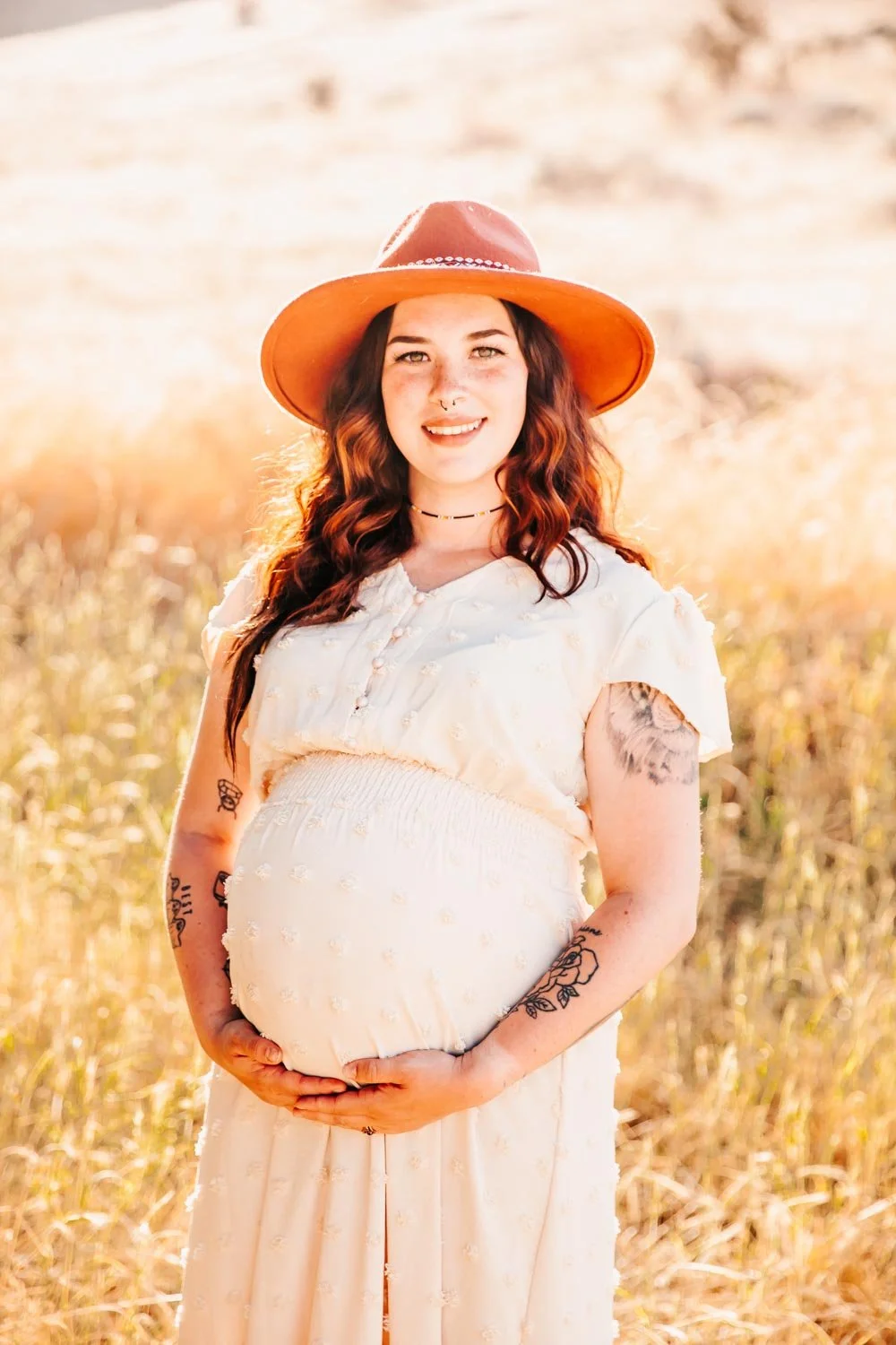 Pregnant woman wearing a hat in a field, smiling and holding her belly.