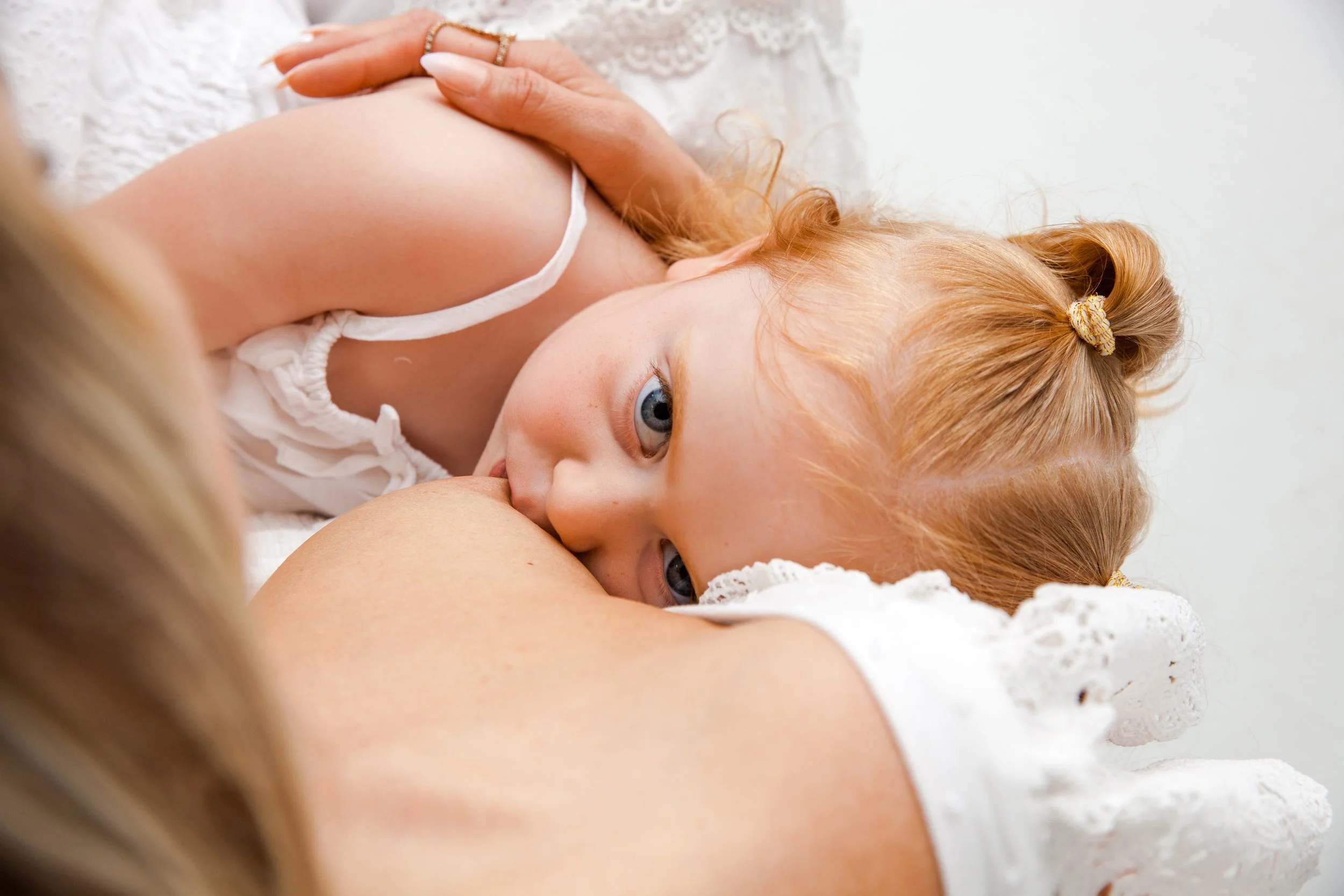 A young child breastfeeding, wearing a white outfit, and being held by an adult in a white garment.