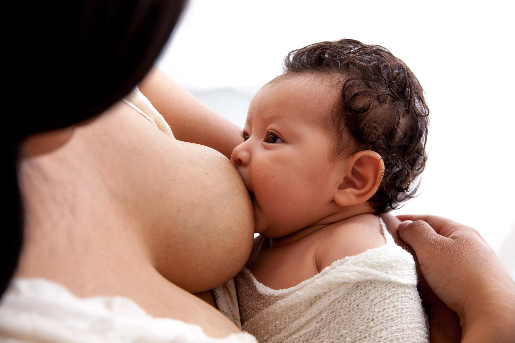 A mother breastfeeding her baby, with the infant wrapped in a white blanket.