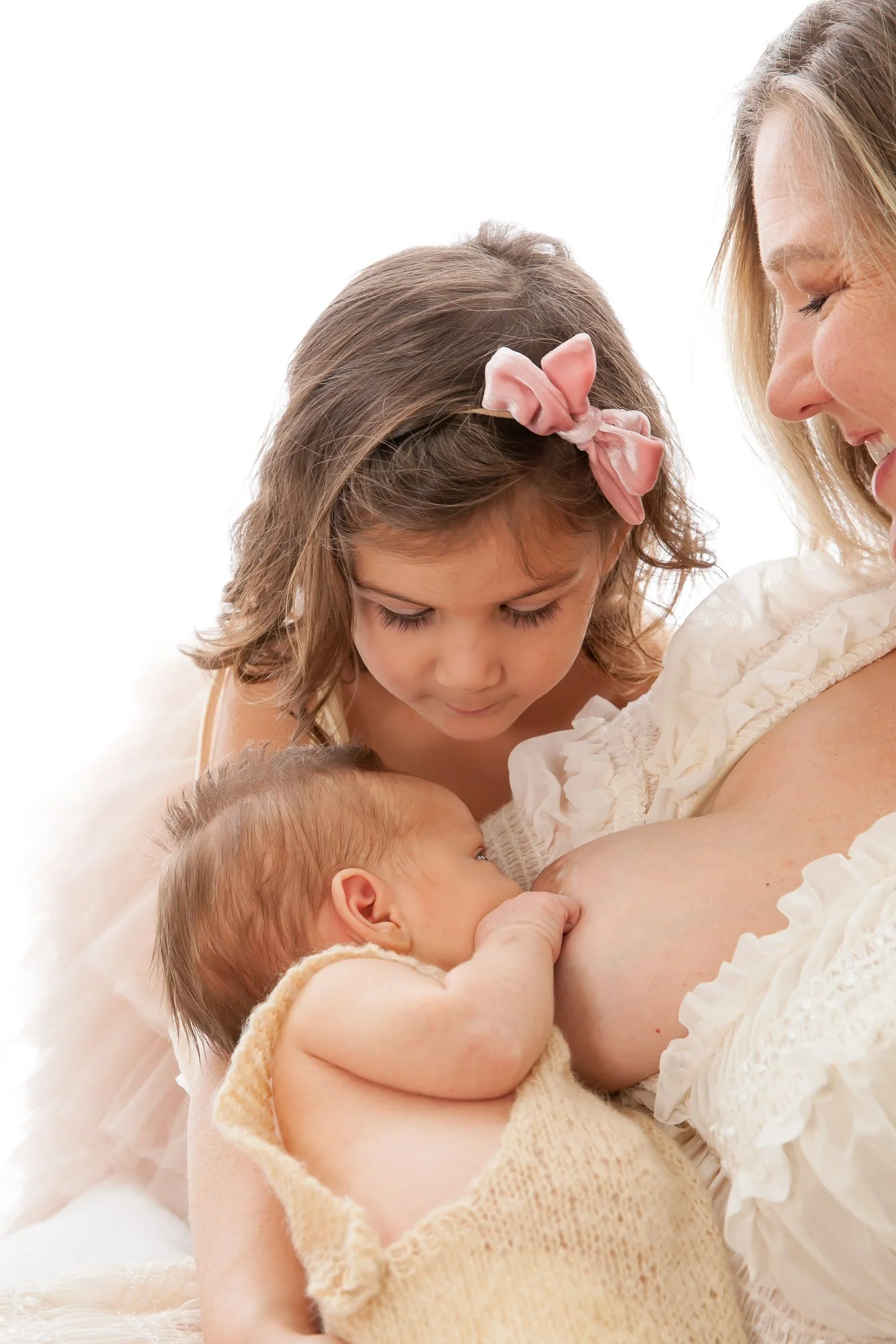 A woman breastfeeding a baby while a young girl with a pink bow looks on.