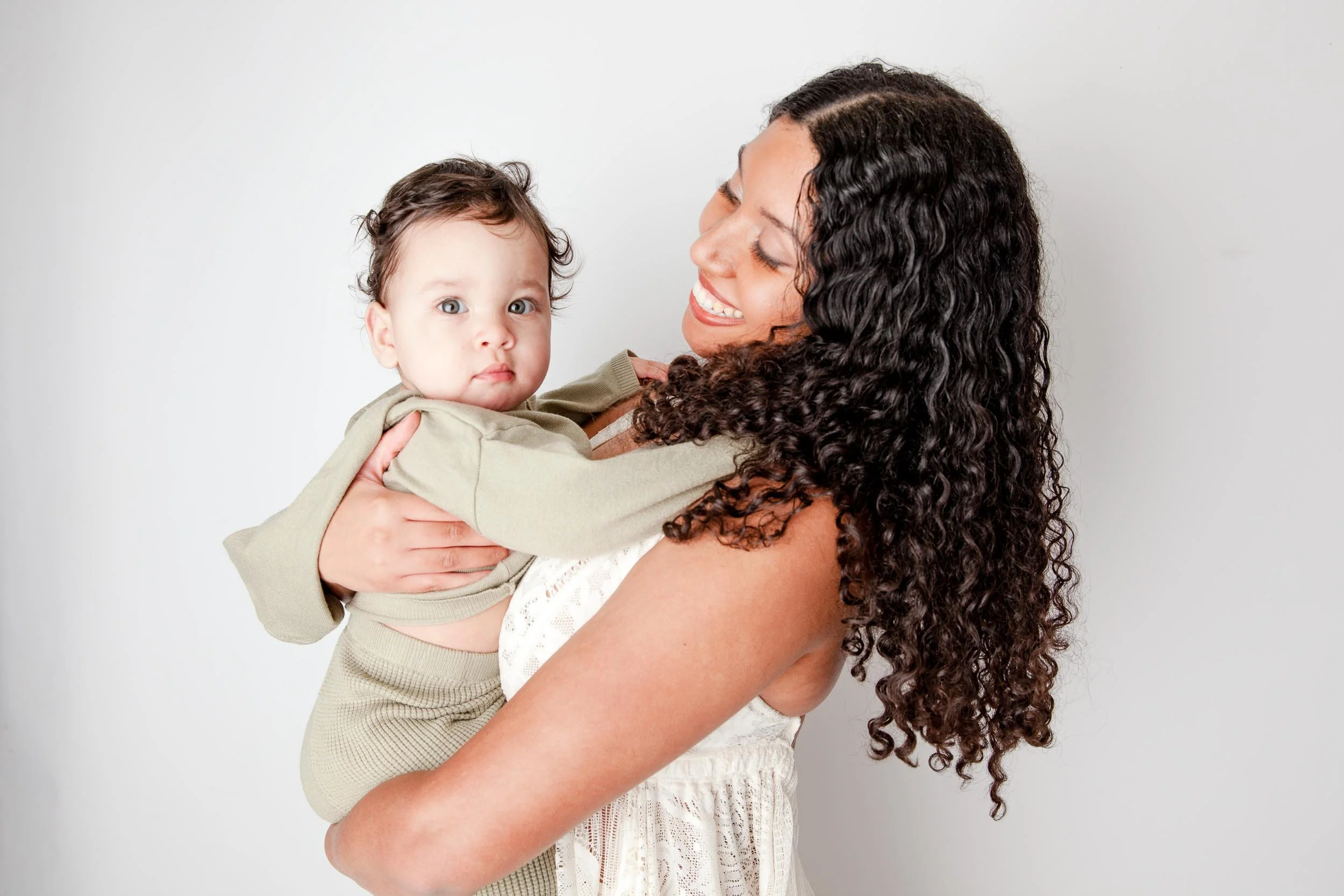 Woman smiling while holding a baby in a light green outfit against a white background.