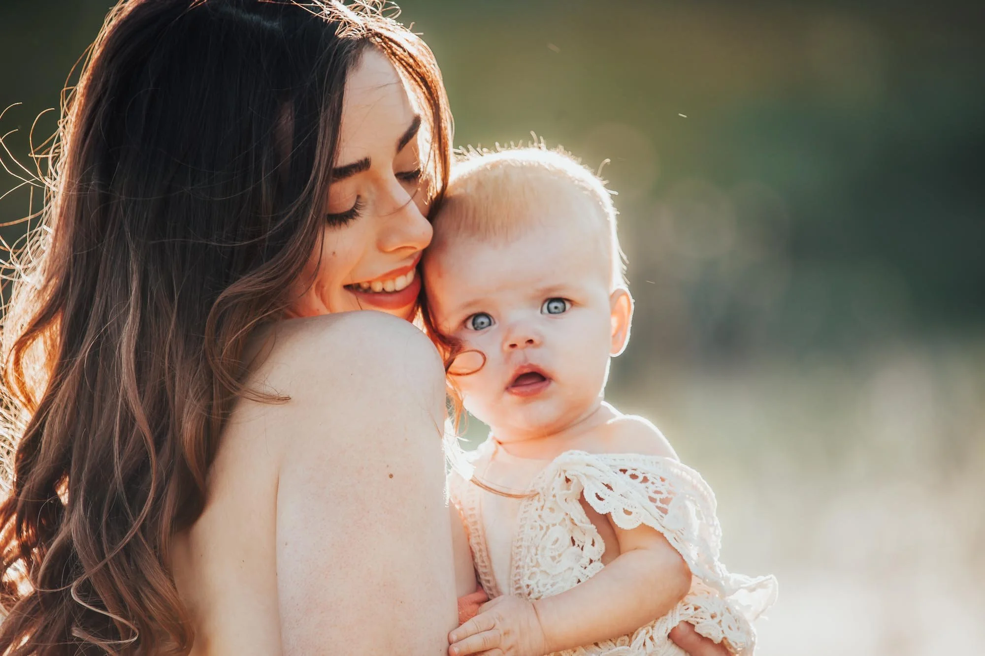 A woman smiling while holding a baby outdoors, with soft lighting highlighting their faces.