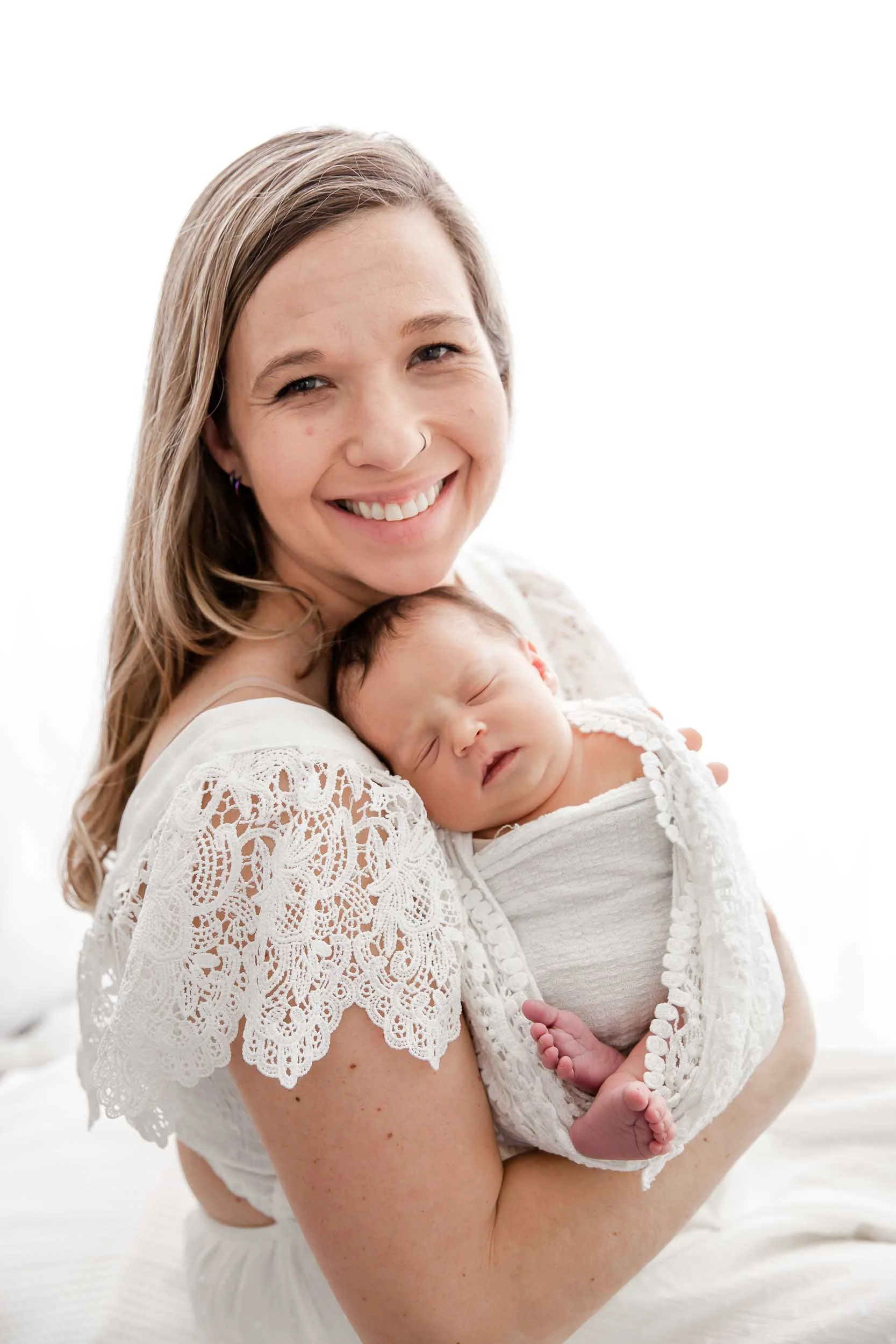 Mother holding newborn baby wrapped in a white blanket, both smiling and wearing white attire, with a plain white background.