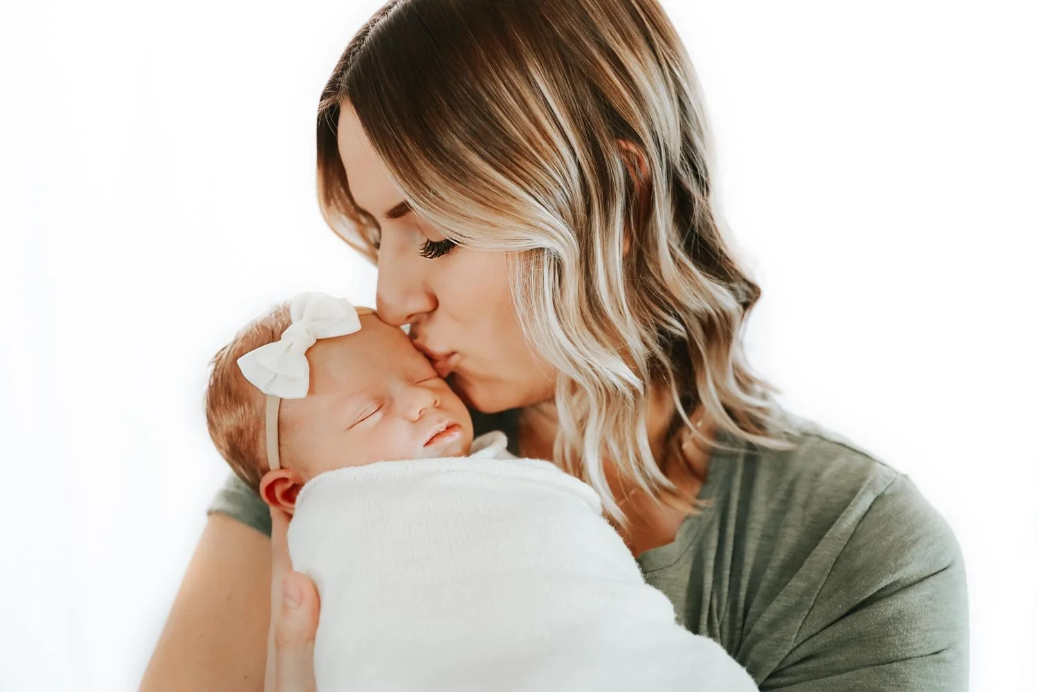 Woman kissing a swaddled newborn baby wearing a white bow.