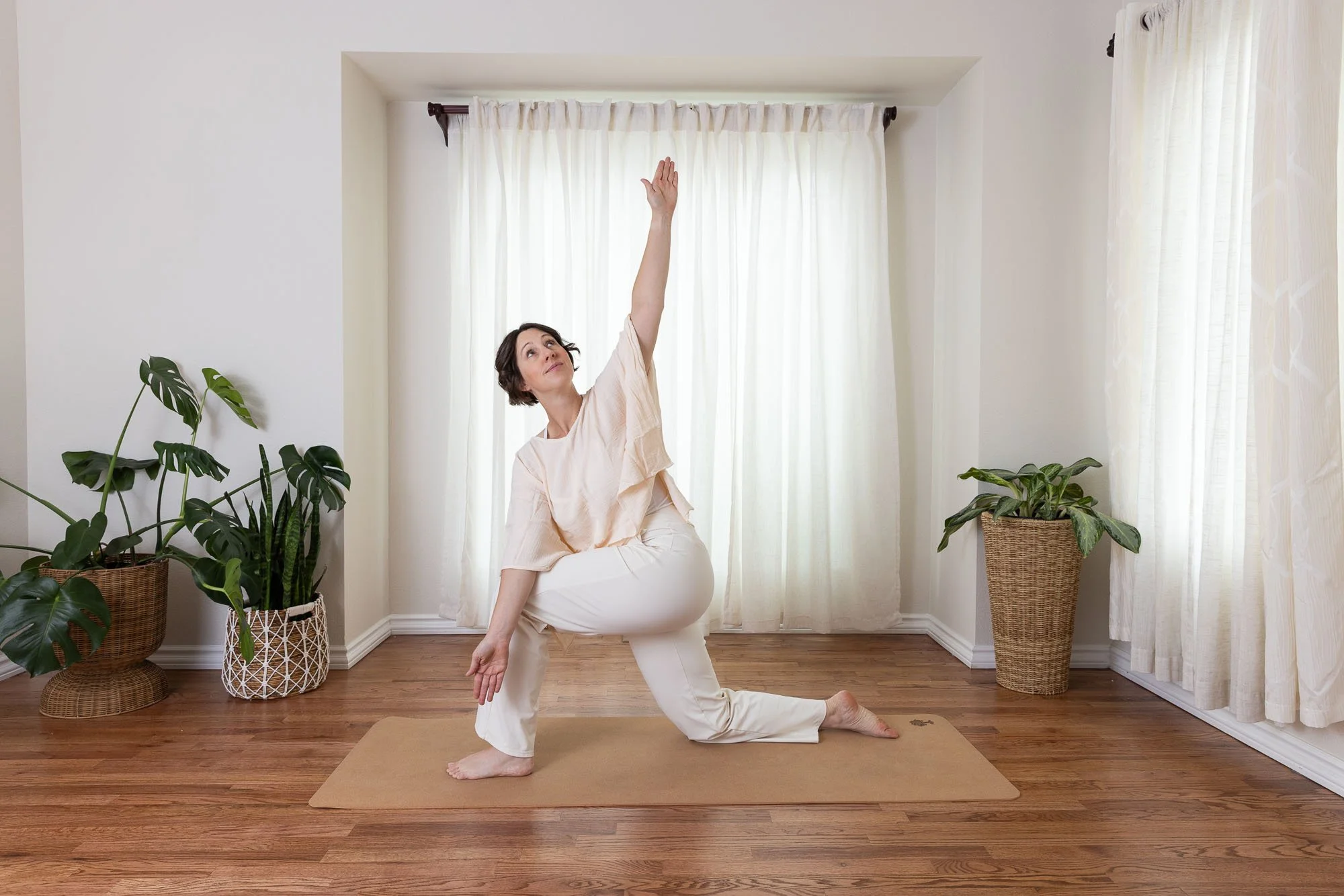 Image of a woman doing yoga in a living room with a big window with sheer curtains and potted plants.